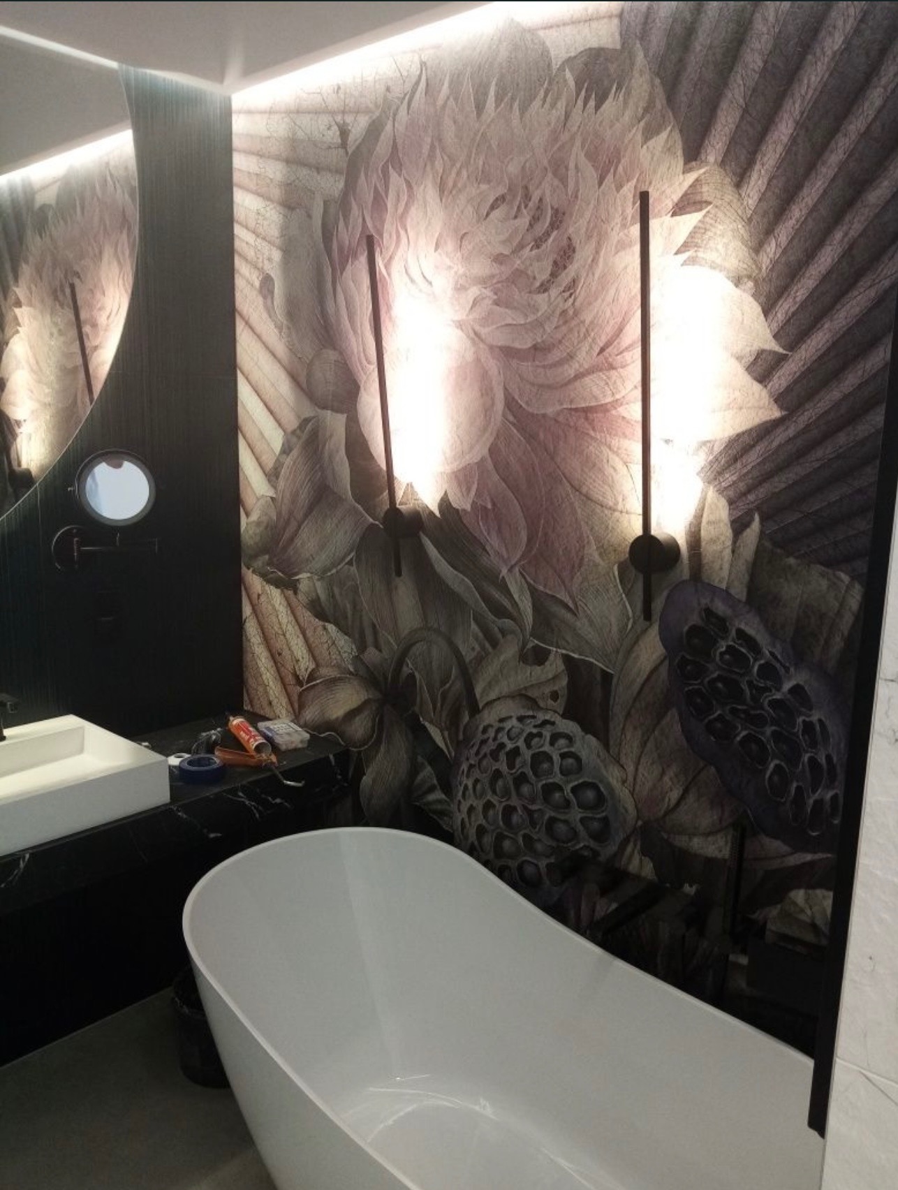 Nowoczesna łazienka z wolnostojącą wanną i umywalką na czarnym blacie, ozdobiona tapetą w kwiaty i oświetleniem punktowym. Widoczne akcesoria montażowe.