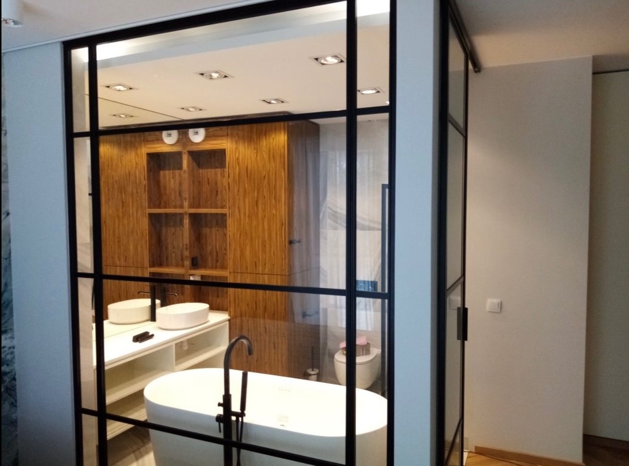 Nowoczesna łazienka za szklaną ścianą z czarnymi szprosami. Wolnostojąca wanna, umywalki nablatowe i drewniane szafki tworzą eleganckie wnętrze.