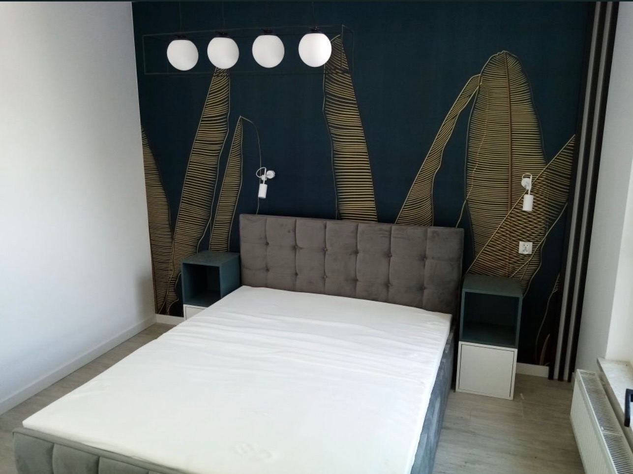 Sypialnia z łóżkiem, szafkami nocnymi i dekoracyjną ścianą w ciemnych barwach z motywem liści. Nad łóżkiem lampa z kulistymi kloszami.