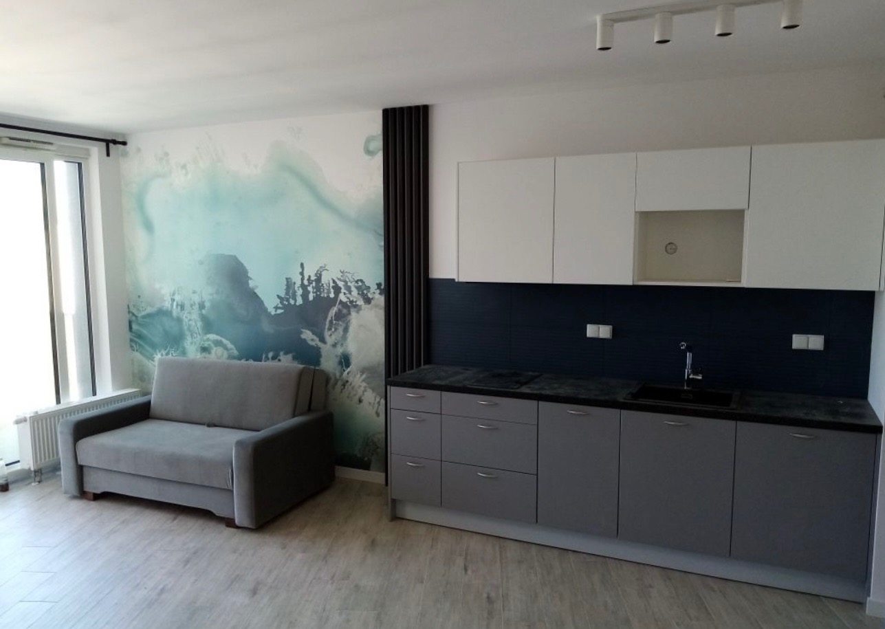 Nowoczesne wnętrze: szara sofa na tle ściany z abstrakcyjnym wzorem, obok zabudowa kuchenna z białymi szafkami górnymi i szarymi dolnymi, czarny panel nad blatem.