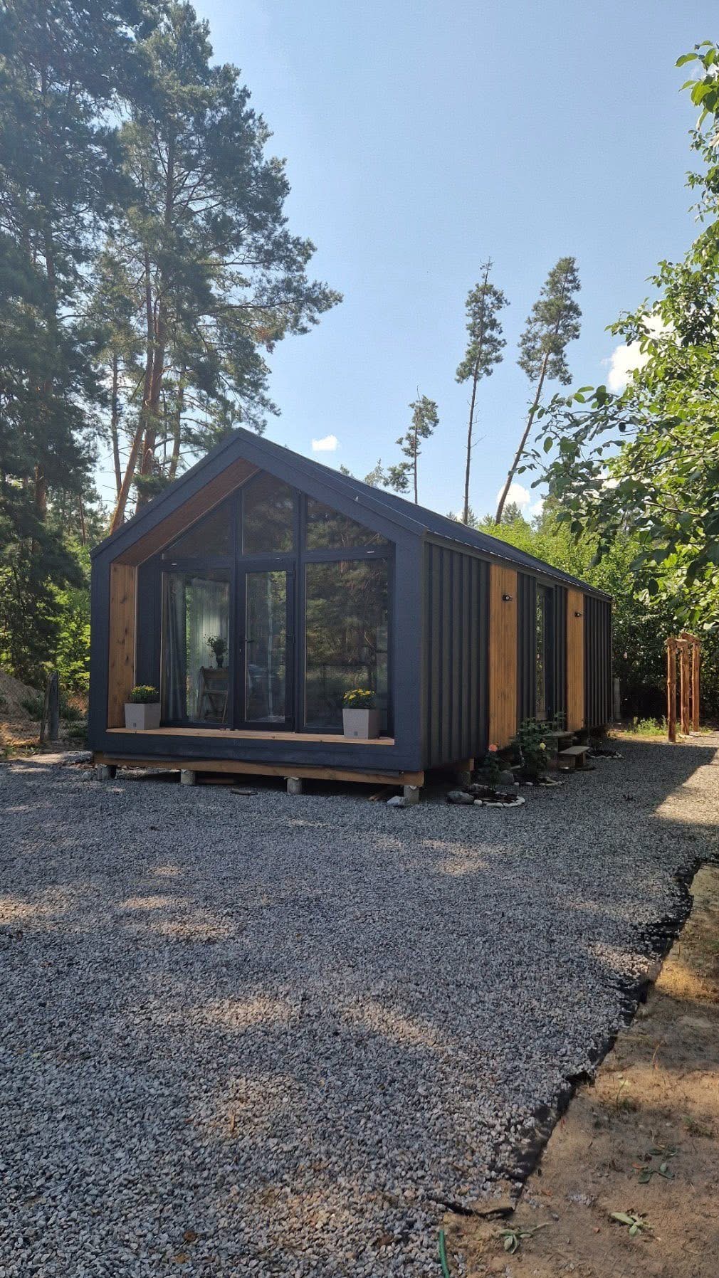 Nowoczesny, parterowy dom modułowy z dużymi przeszkleniami, otoczony zielenią i wysypany żwirem. Widoczne drewniane elementy wykończeniowe.