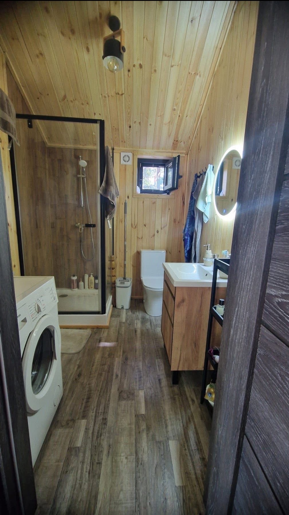 Wykończona łazienka w drewnie: kabina prysznicowa, pralka, umywalka z szafką, toaleta, okrągłe lustro z podświetleniem. Całość w jasnym drewnie, z ciemniejszymi akcentami.