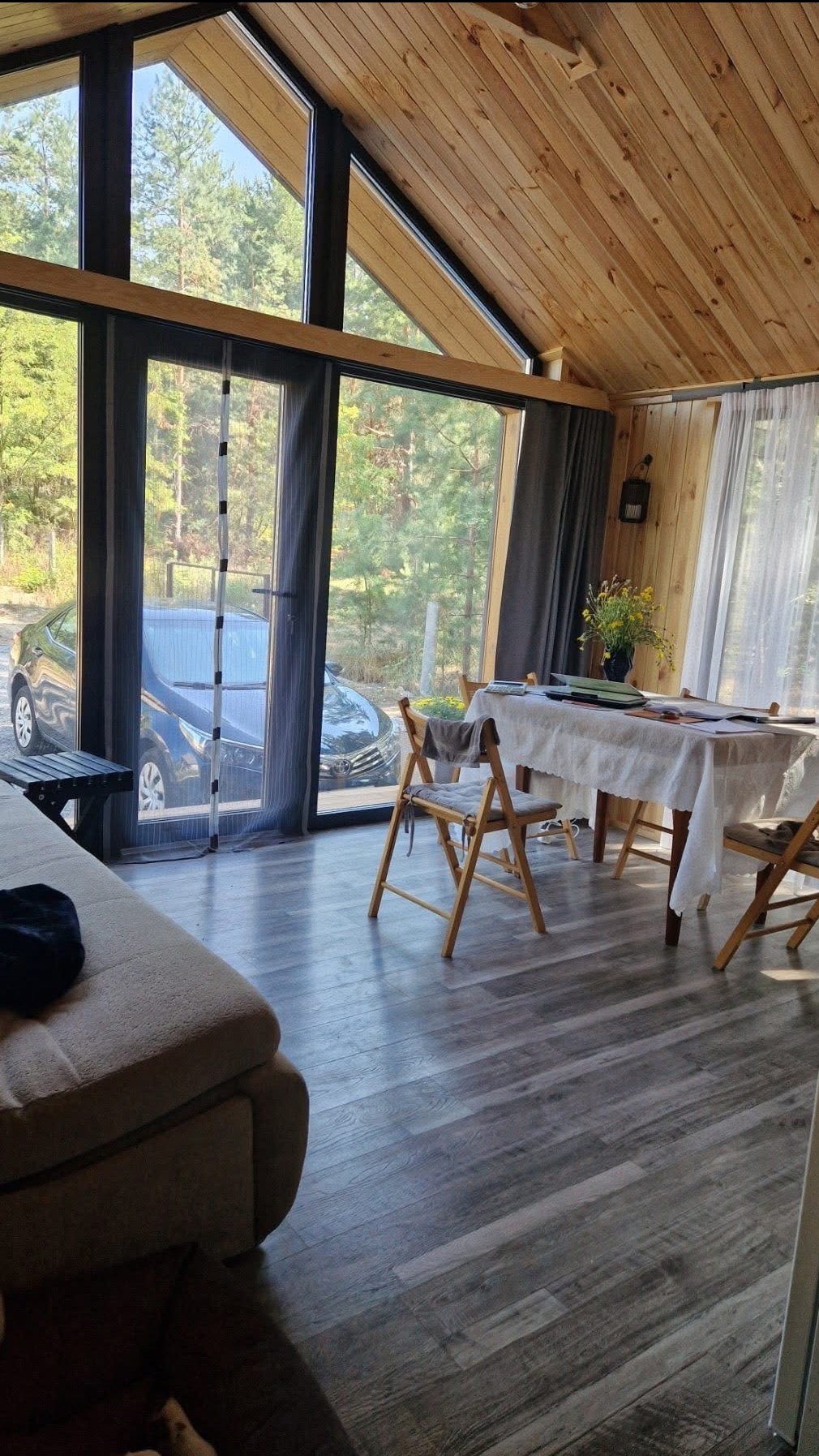 Jasne wnętrze z drewnianym sufitem i dużym oknem z widokiem na zieleń. Wnętrze urządzone w stylu skandynawskim, z drewnianymi krzesłami i stołem nakrytym białym obrusem.