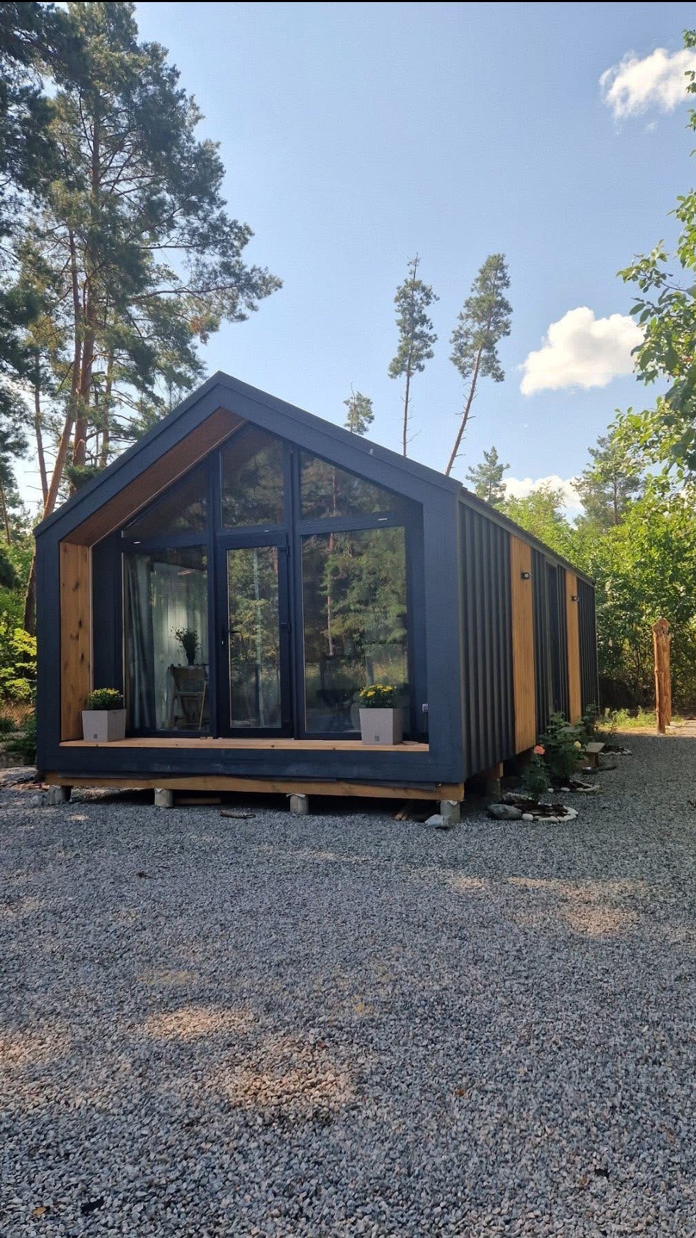 Nowoczesny, modułowy domek letniskowy z dużymi przeszkleniami, otoczony zielenią i wysypany żwirem. Widoczne detale wykończenia drewnianego i donice z kwiatami.