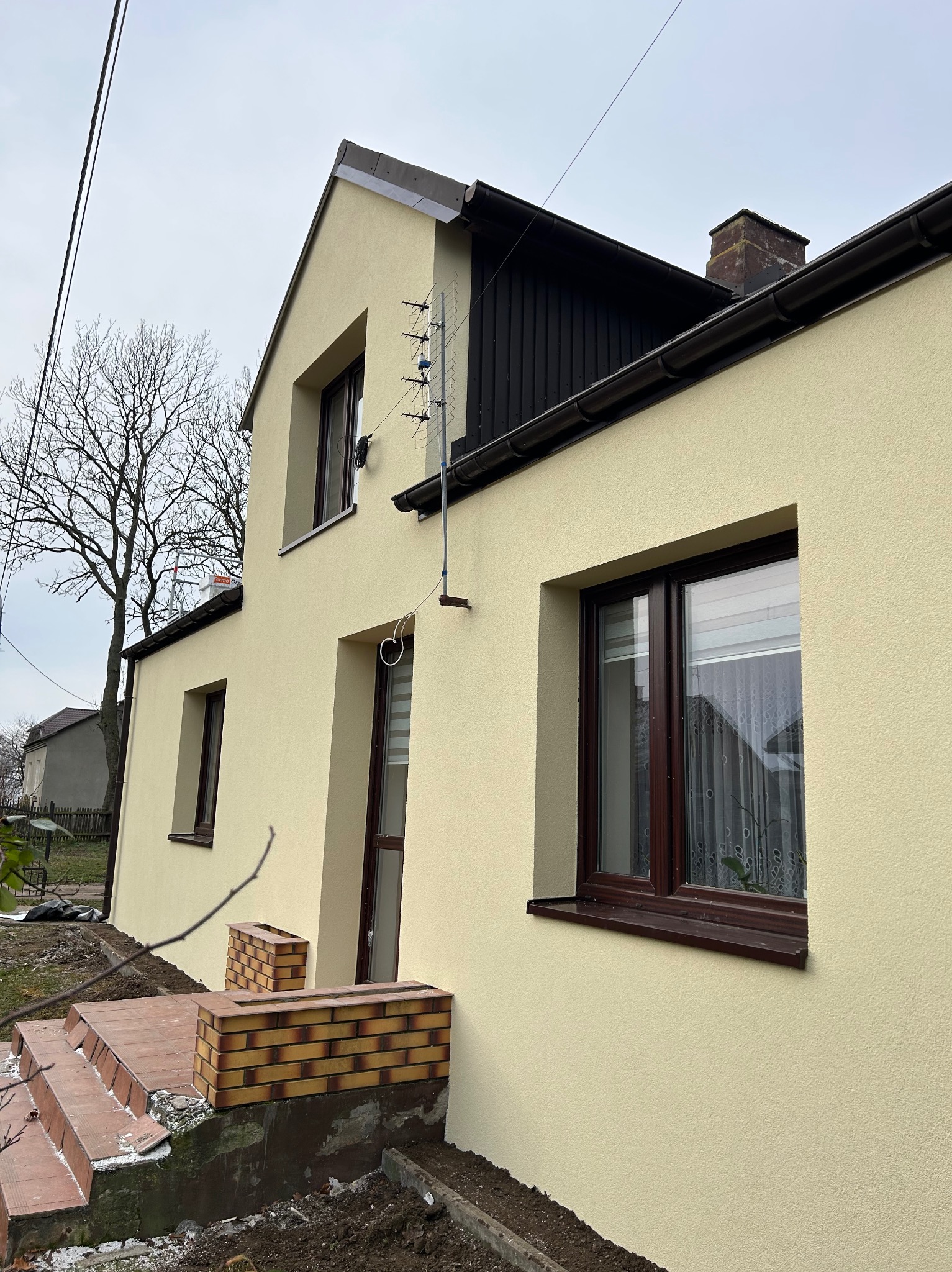 Jasna elewacja dwukondygnacyjnego domu z brązowymi oknami i wykończeniem dachu. Widoczna antena satelitarna i fragment schodów wejściowych z cegły.