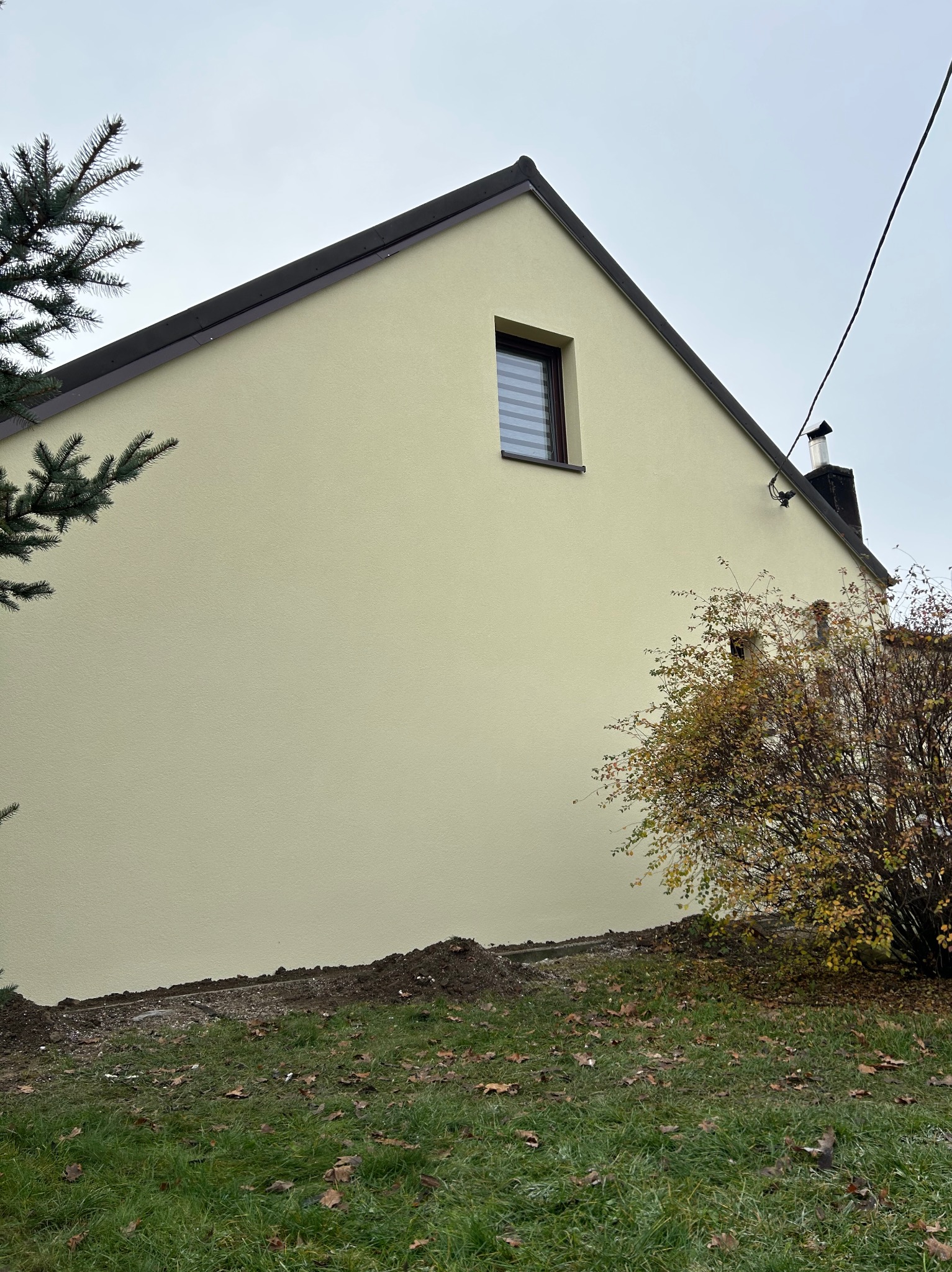 Jasna elewacja domu jednorodzinnego z małym oknem, widoczna z boku, z fragmentem zieleni i drzewa iglastego. Świeżo wykończona, gładka powierzchnia.