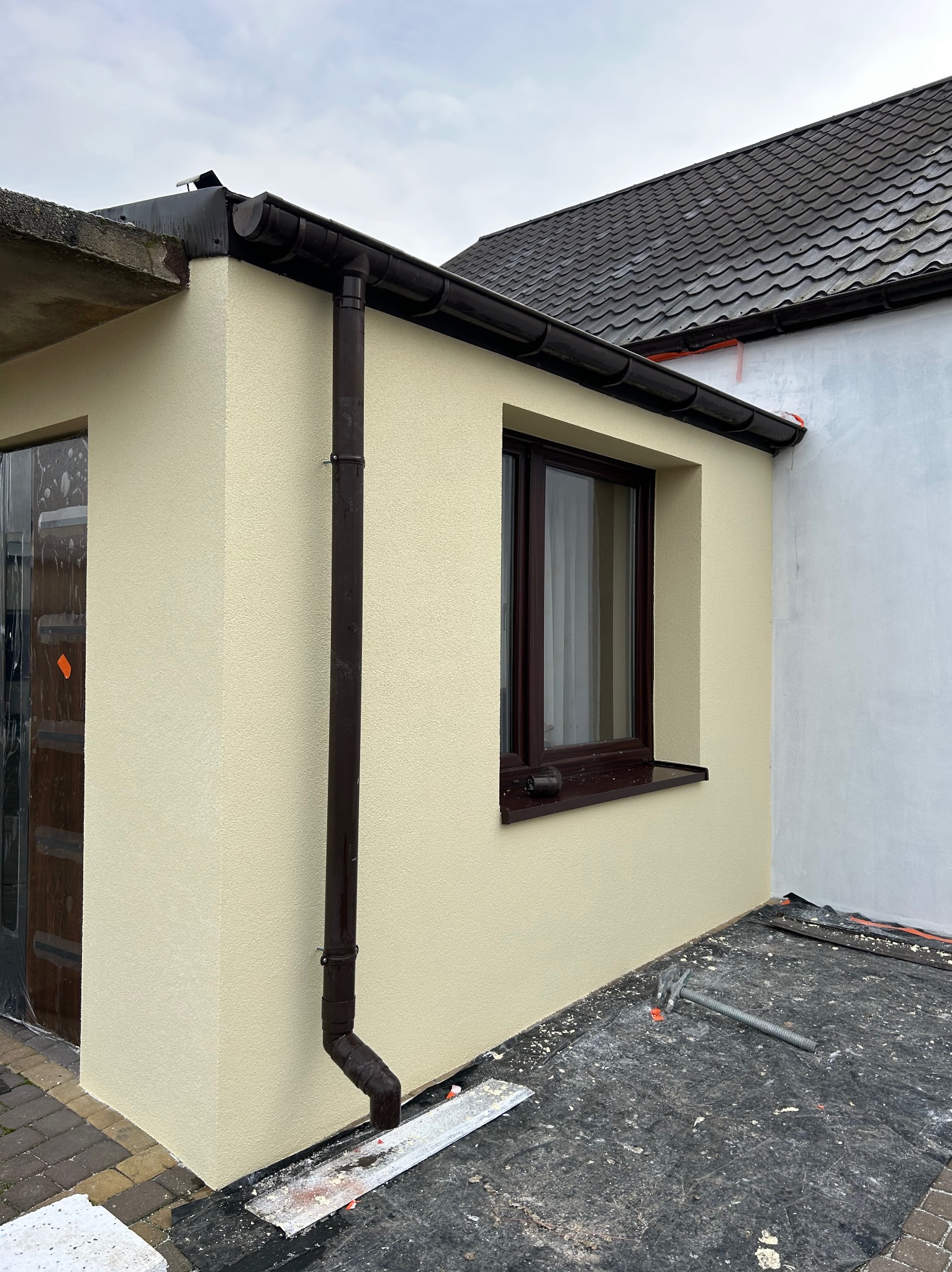 Jasna elewacja budynku z brązową rynną i oknem. Widoczne narzędzia i zabezpieczenia na podłożu, sugerujące prace wykończeniowe. Część budynku pomalowana na biało.