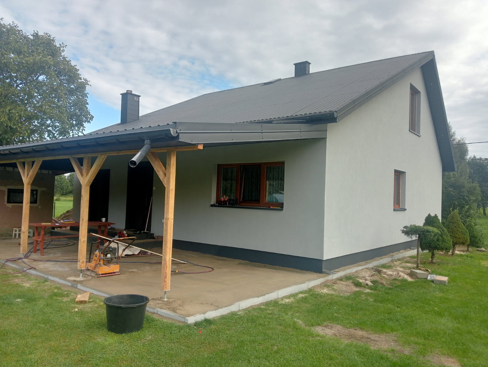 Dom po tynkowaniu elewacji na szaro, z drewnianą wiatą i widocznym sprzętem budowlanym. Nowe okna z brązowymi ramami. Widok z zewnątrz na świeżo wykończony budynek.