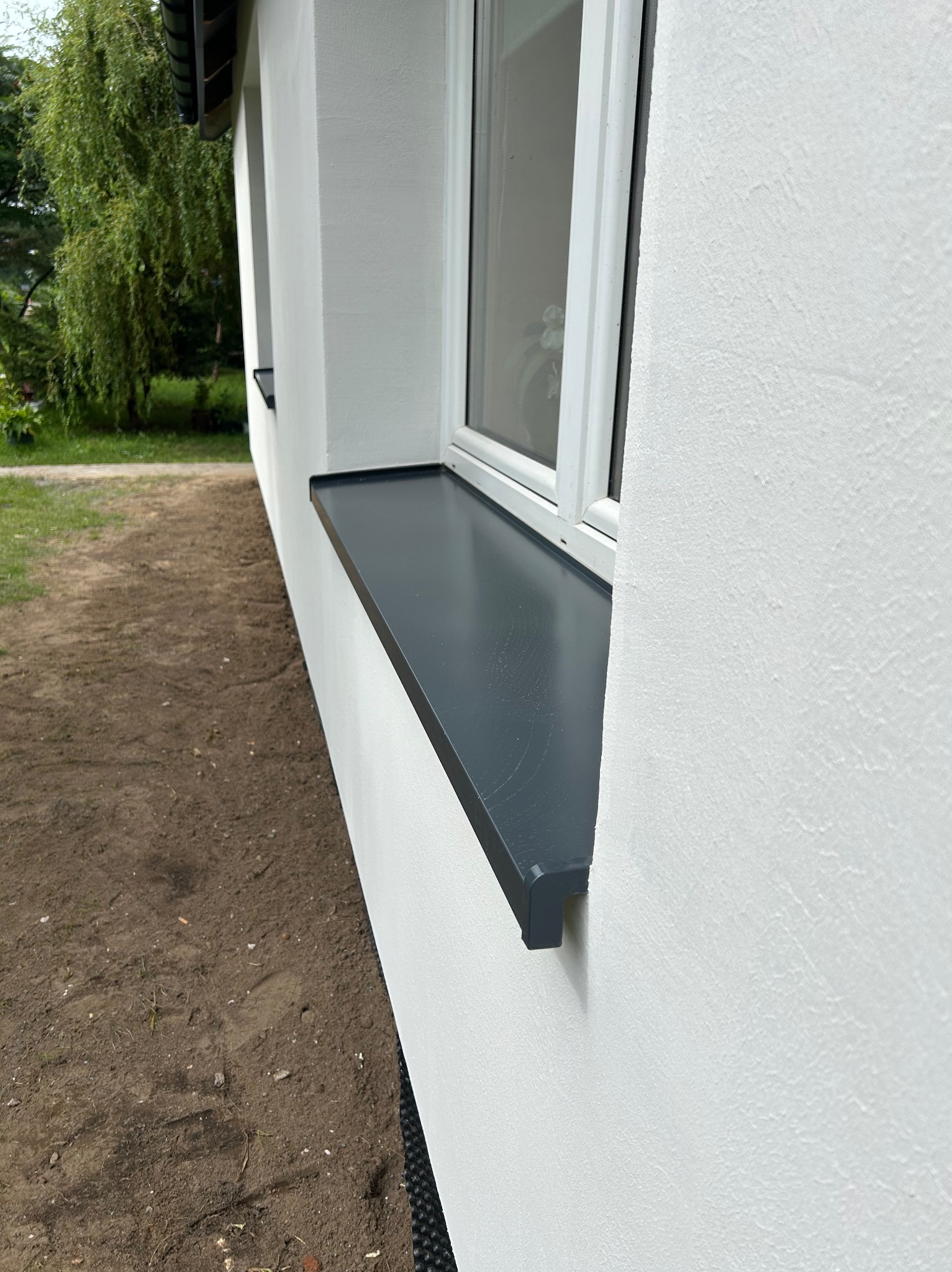 Biała elewacja budynku z zamontowanym, grafitowym parapetem zewnętrznym. Widoczna faktura tynku i fragment trawnika. Ujęcie z dołu, podkreślające detale wykończenia.