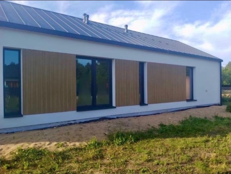 Nowoczesny dom jednorodzinny z jasną elewacją i drewnianymi panelami. Widoczne okna z czarnymi ramami i ciemny dach. Ujęcie z poziomu gruntu, trawa i piasek na pierwszym planie.