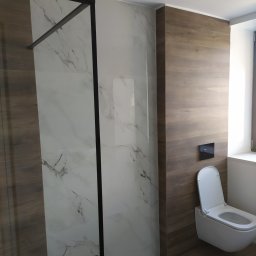 Fili-Fliz - Nowoczesna łazienka z kabiną typu walk-in, wykończona płytkami imitującymi drewno i marmur. Uwagę zwraca minimalistyczna toaleta podwieszana i czarna rama kabiny.