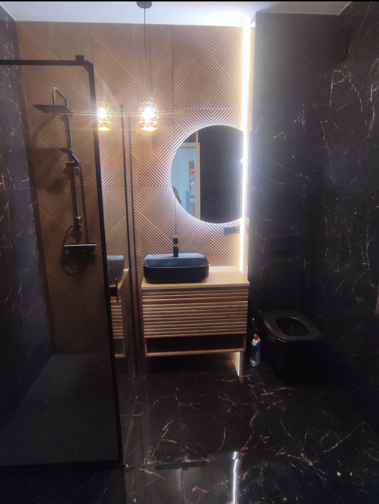 Nowoczesna łazienka z czarnymi marmurowymi płytkami, drewnianą szafką pod umywalką i okrągłym lustrem z podświetleniem LED. Kabina prysznicowa ze szklanymi drzwiami.