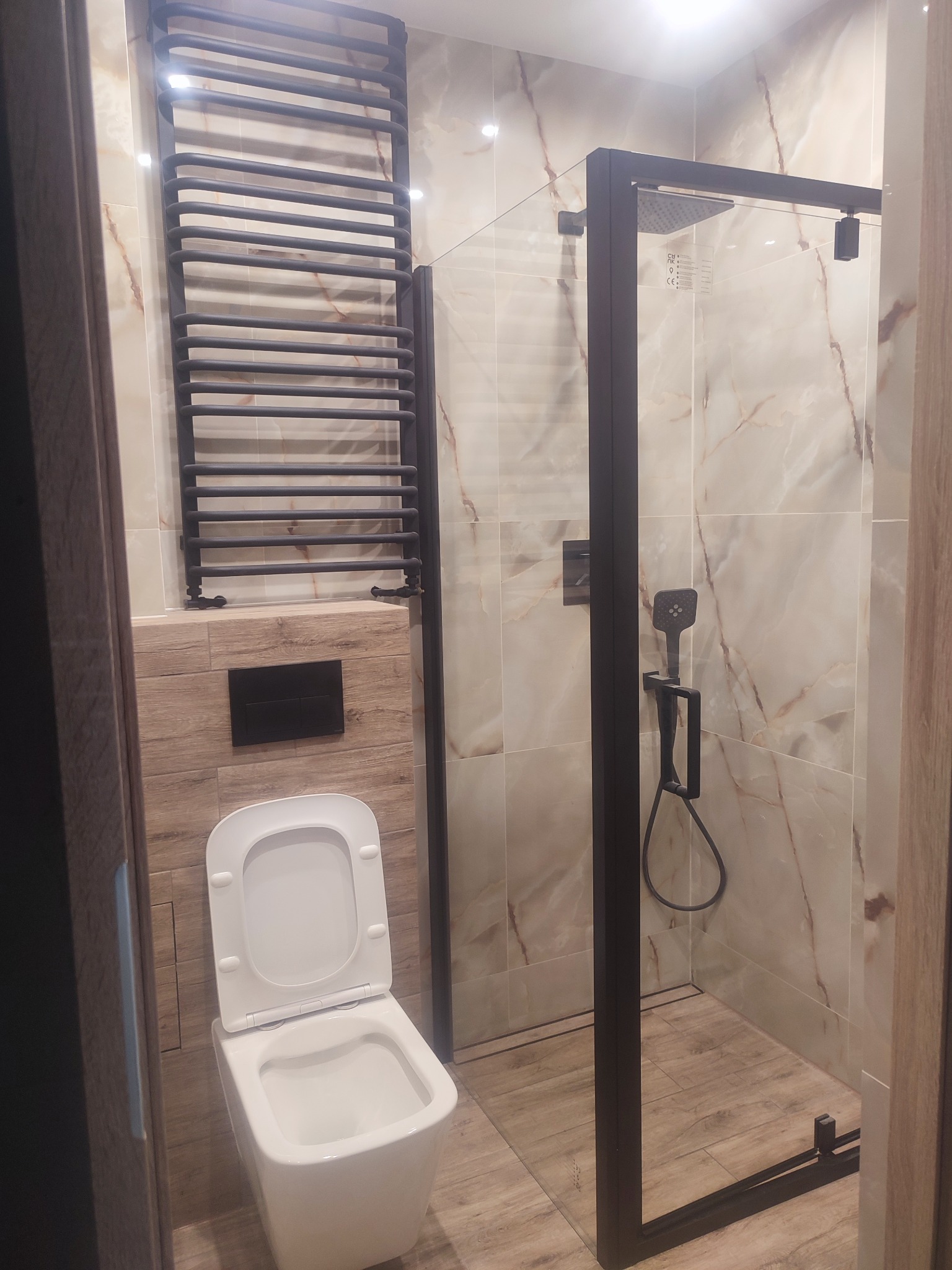 Nowoczesna łazienka z kabiną prysznicową typu walk-in z czarnymi profilami, wiszącą toaletą i grzejnikiem drabinkowym w ciemnym kolorze na tle marmurowych płytek.