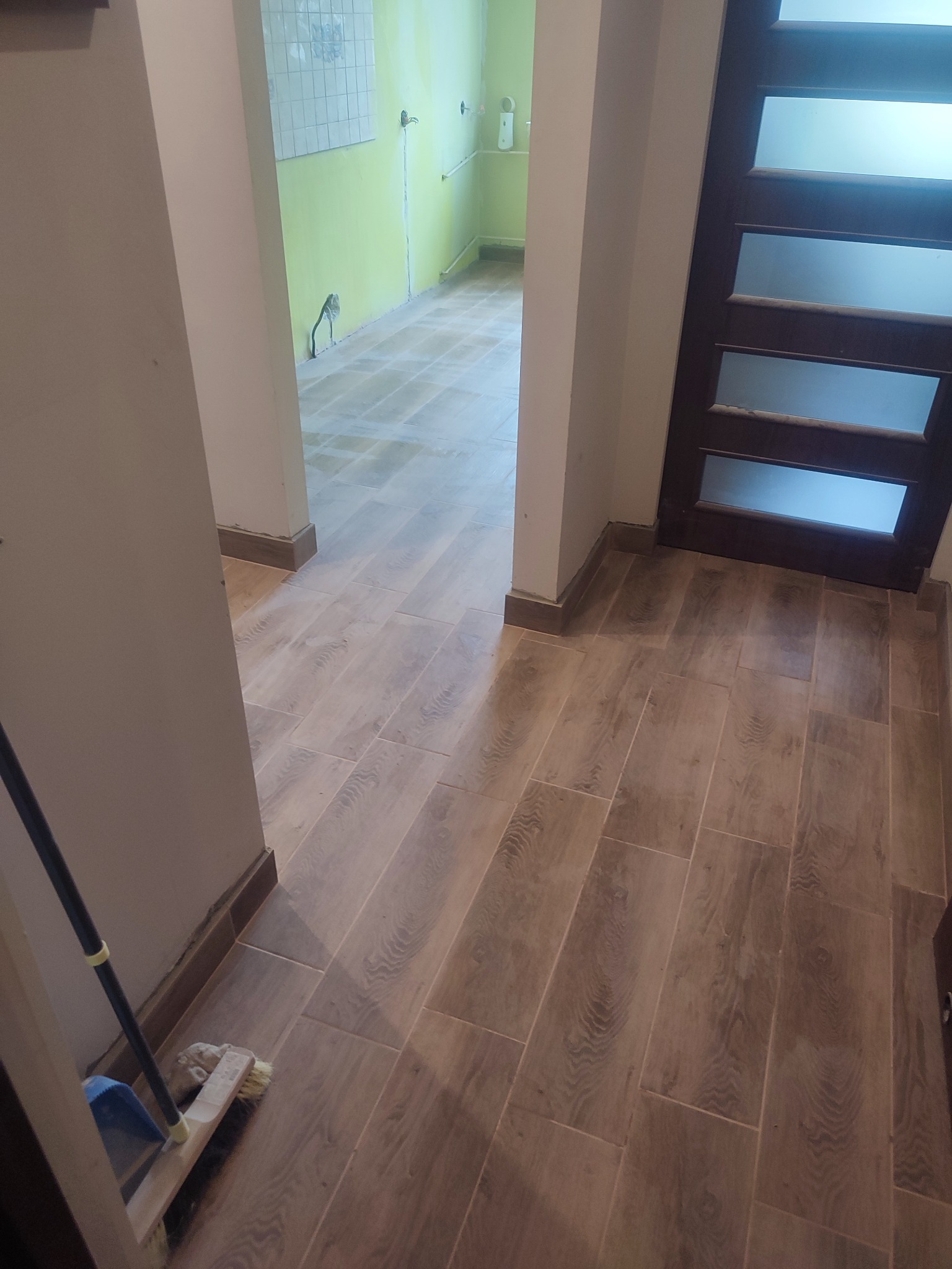 Ułożona podłoga z płytek imitujących drewno w korytarzu. Widoczne fragmenty ścian w kolorze beżowym i zielonym. W tle łazienka z białymi płytkami.