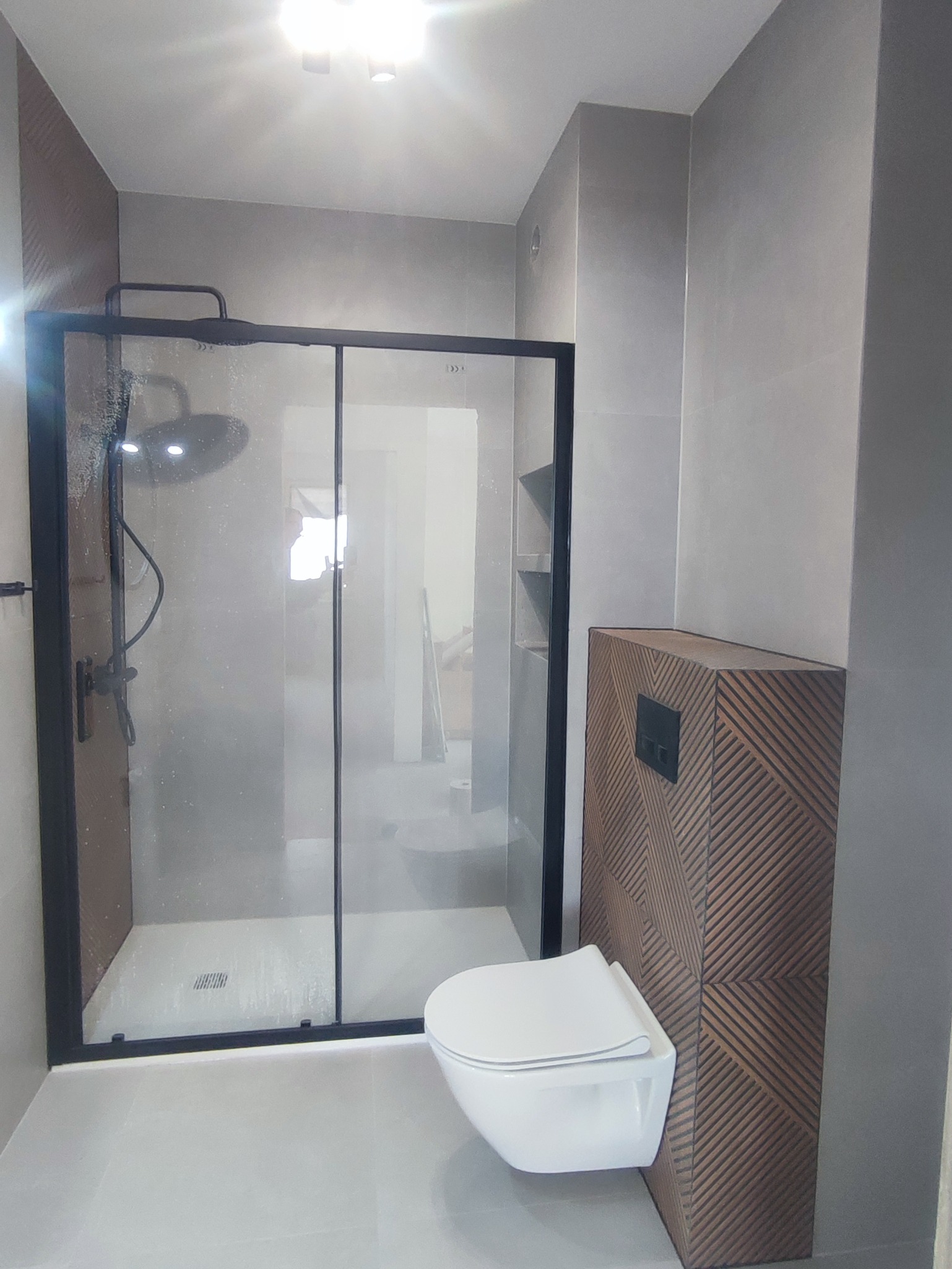 Nowoczesna łazienka po remoncie: kabina prysznicowa typu walk-in z czarnymi ramami, podwieszana toaleta z zabudową w dekoracyjne, drewniane pasy, szare płytki.