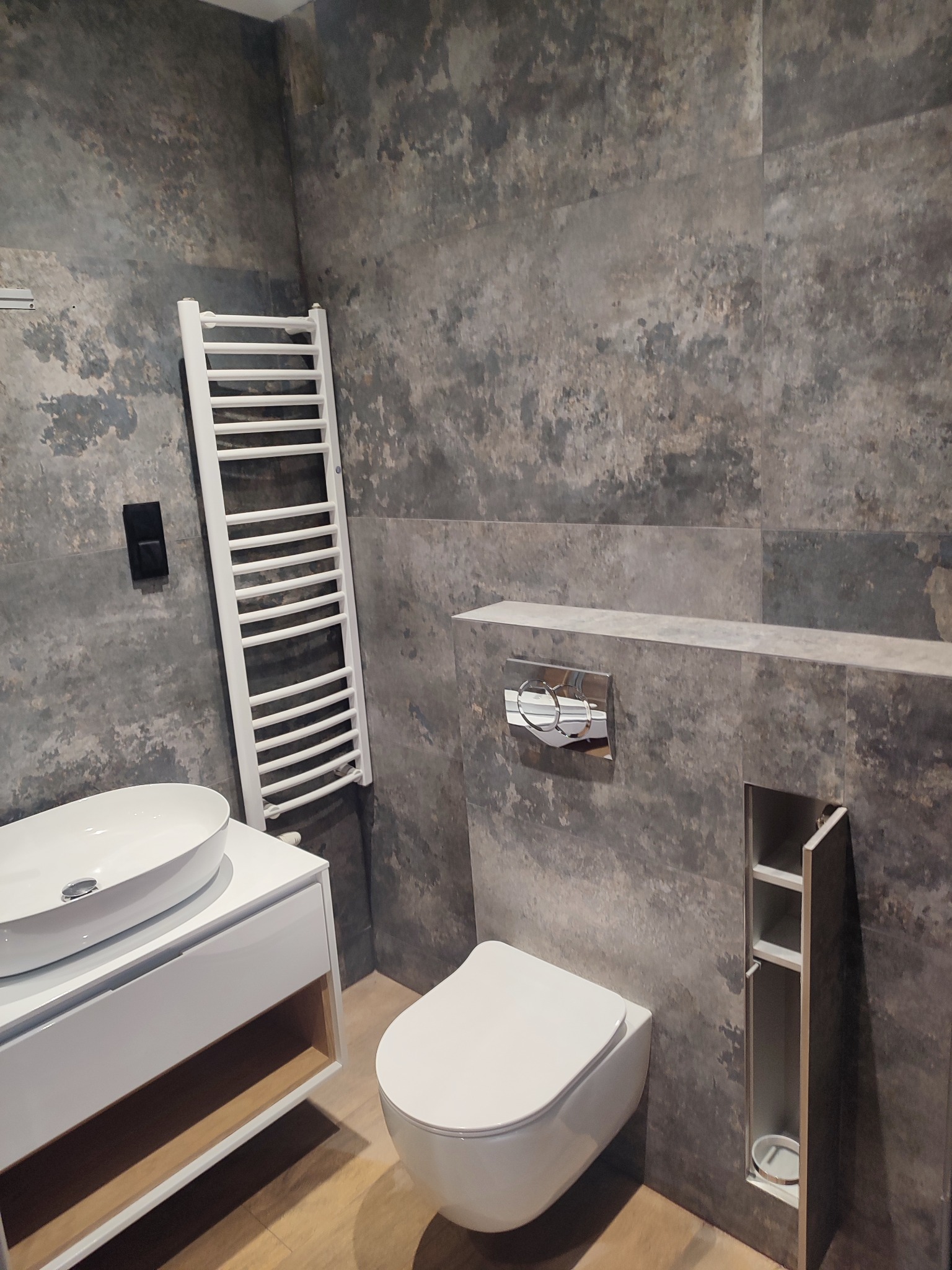 Nowoczesna łazienka z szarymi płytkami imitującymi beton, białą umywalką nablatową i podwieszaną toaletą. Grzejnik drabinkowy i ukryta szafka w ścianie.