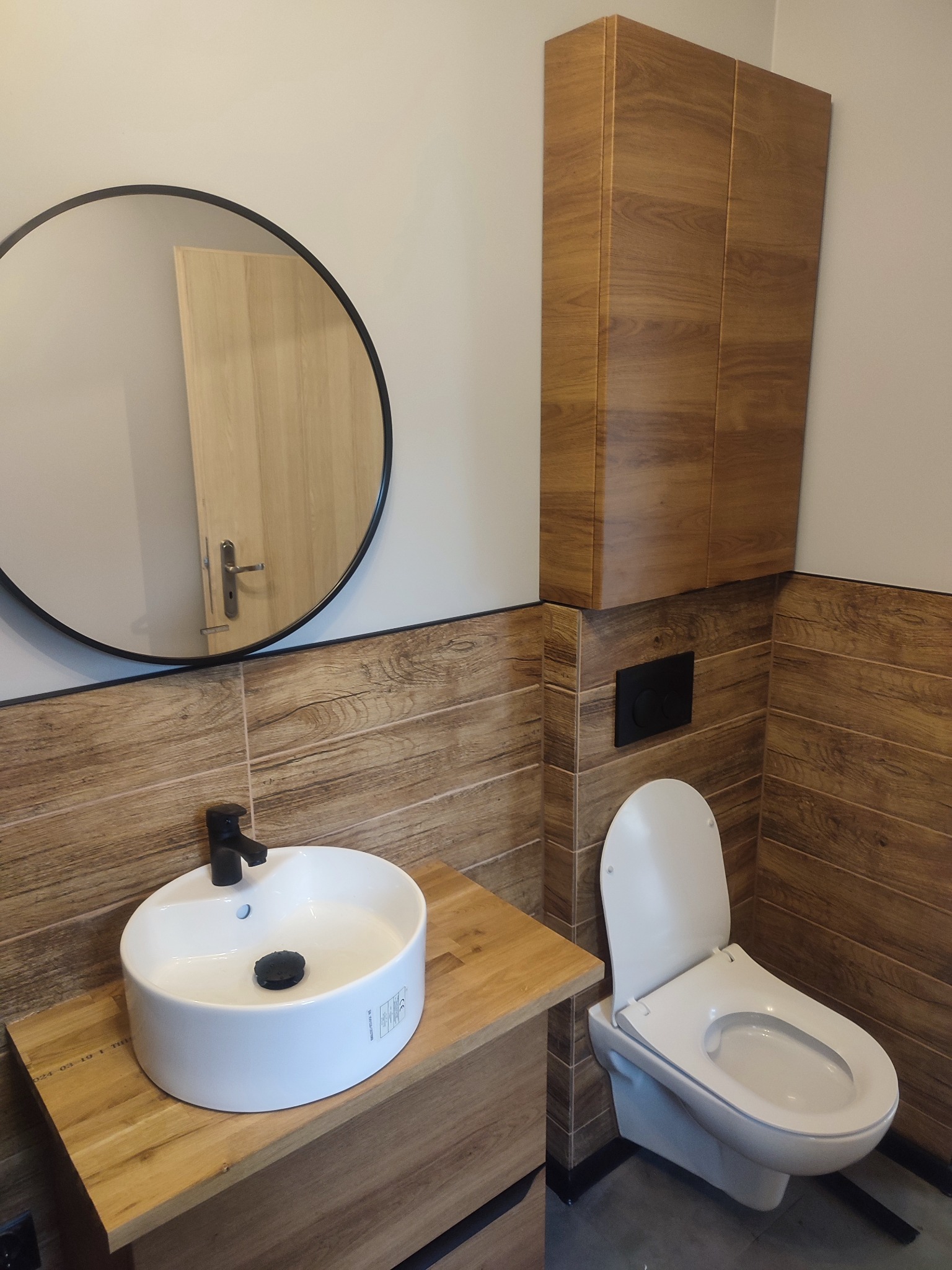 Nowoczesna łazienka z drewnopodobnymi płytkami, okrągłym lustrem i białą umywalką nablatową z czarną armaturą. Wisząca szafka nad toaletą.