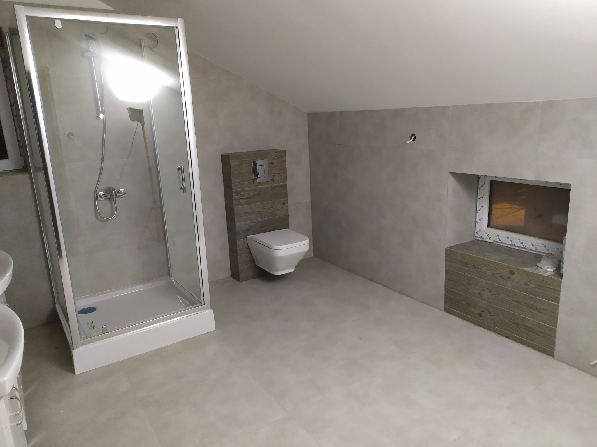 Wykończona łazienka na poddaszu w Bukownie. Kabina prysznicowa, toaleta podwieszana i obudowa okna w drewnopodobnych płytkach. Jasne płytki na ścianach i podłodze.