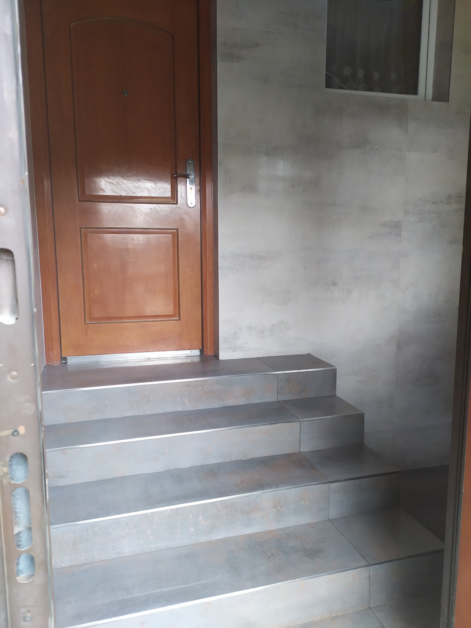 Wykończone schody wejściowe z szarych płytek, prowadzące do brązowych drzwi. Ściany wyłożone płytkami w odcieniach szarości. Widoczne okno w górnej części ściany.