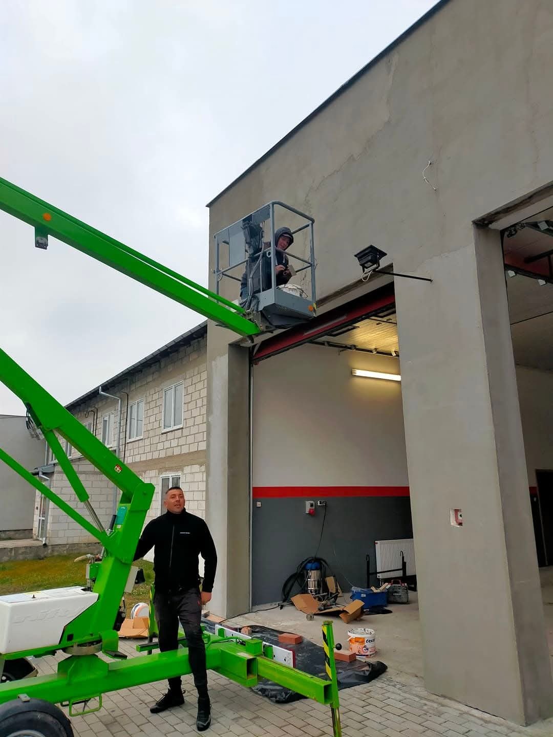 Praca na wysokości: Dwóch mężczyzn na podnośniku koszowym maluje elewację budynku z otwartą bramą garażową. Jeden stoi na ziemi, drugi w koszu podnośnika.