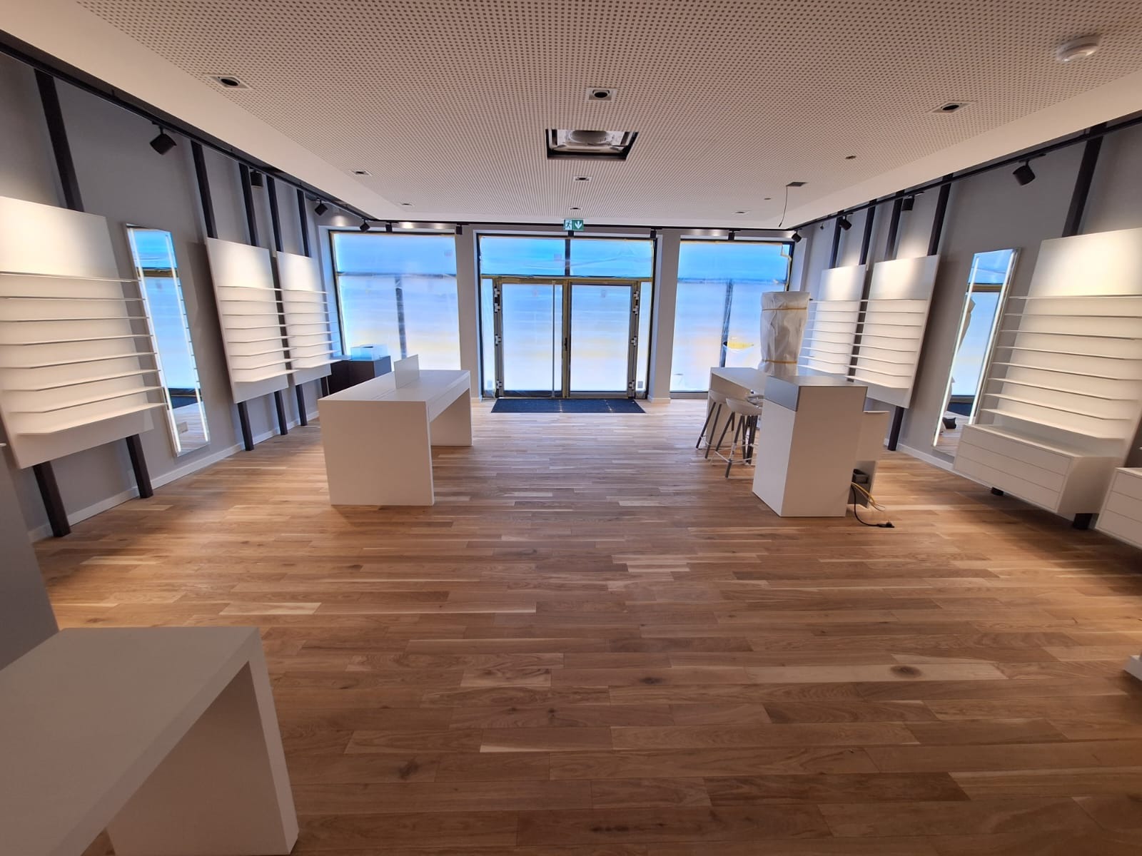 Jasne, przestronne wnętrze sklepu optycznego z drewnianą podłogą, białymi meblami i oświetleniem punktowym. Okna zasłonięte folią ochronną.