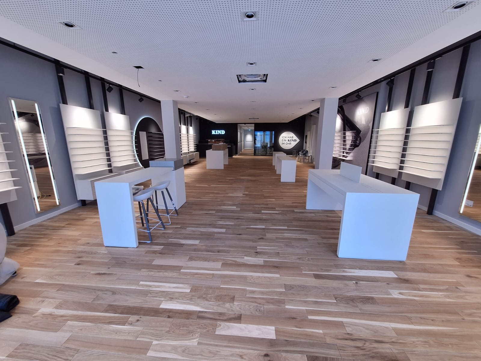 Jasne drewniane panele podłogowe w przestronnym, nowoczesnym salonie optycznym z białymi meblami i szarymi ścianami. Widok w głąb lokalu.