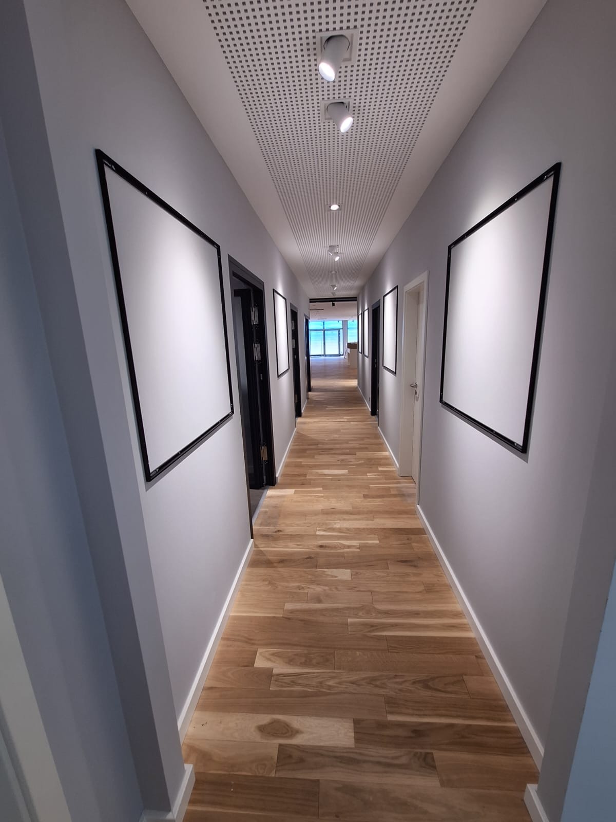 Długi korytarz biurowy z drewnianą podłogą, szarymi ścianami i podwieszanym sufitem z oświetleniem punktowym. Na ścianach wiszą puste ramy.
