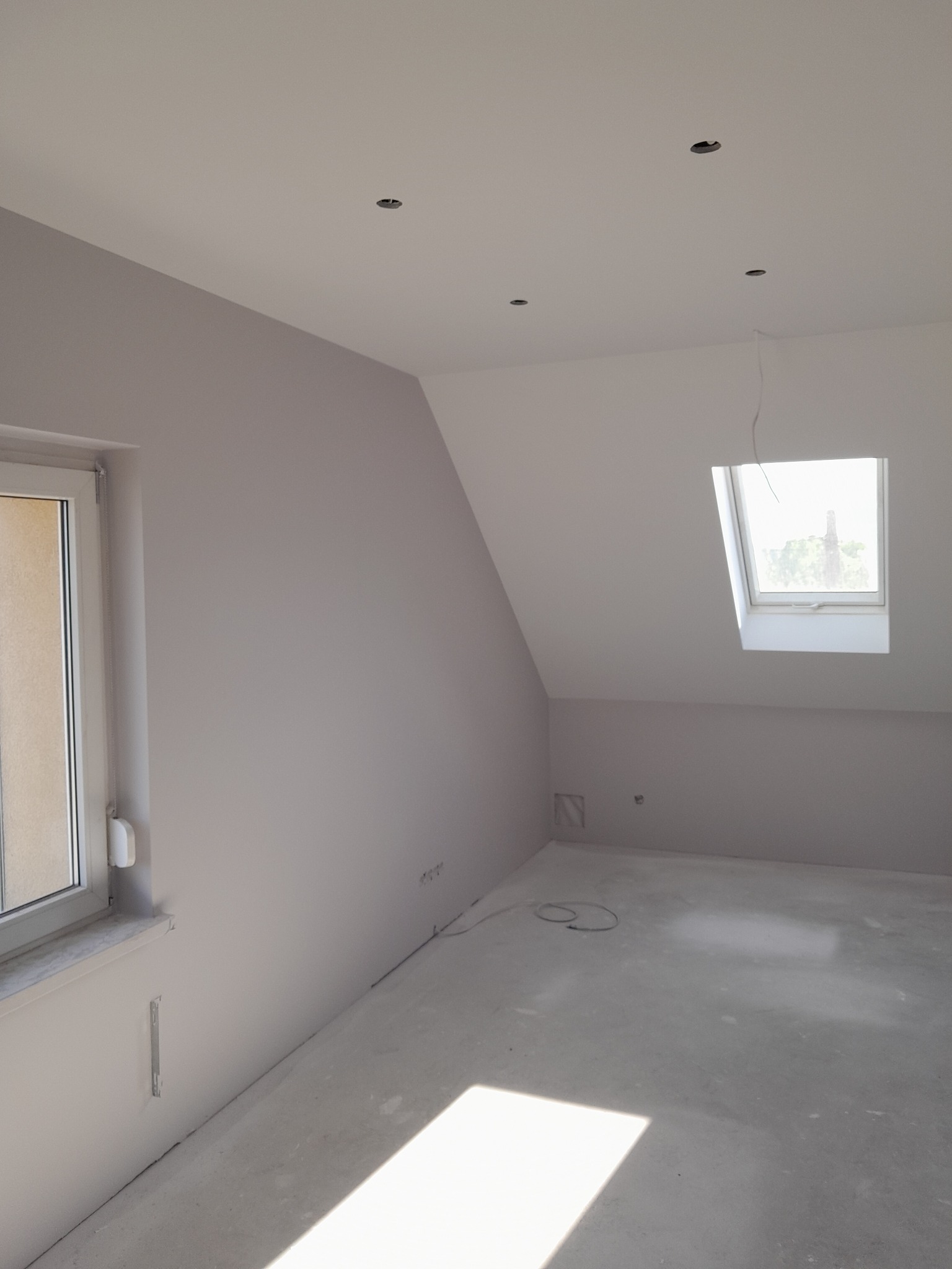 Pomalowane na szaro poddasze ze skośnym sufitem i oknem dachowym. Widoczne gniazdka elektryczne i otwory na oświetlenie w suficie. Podłoga betonowa.