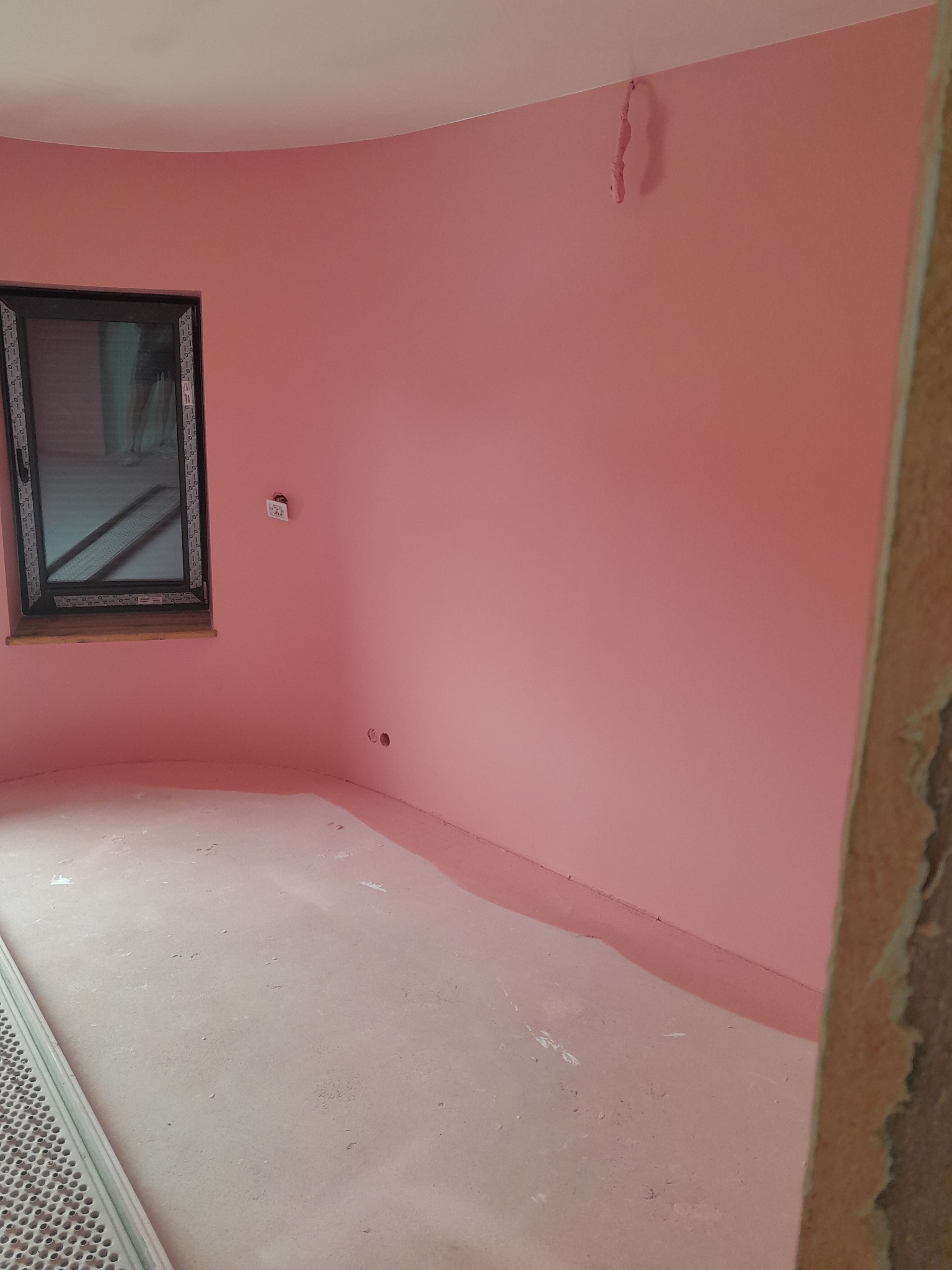 Różowe ściany w okrągłym pokoju z oknem i wystającymi kablami. Świeżo pomalowane wnętrze z widocznymi śladami po remoncie. Minimalistyczny design, nietypowy kształt pomieszczenia.