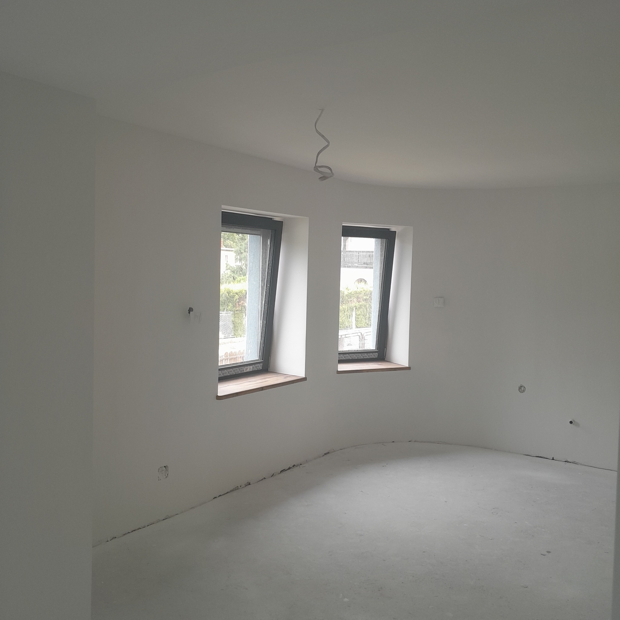 Wnętrze po malowaniu na biało z dwoma oknami. Widoczne ślady po instalacjach elektrycznych i niedoskonałości podłogi. Minimalistyczny, surowy wygląd.