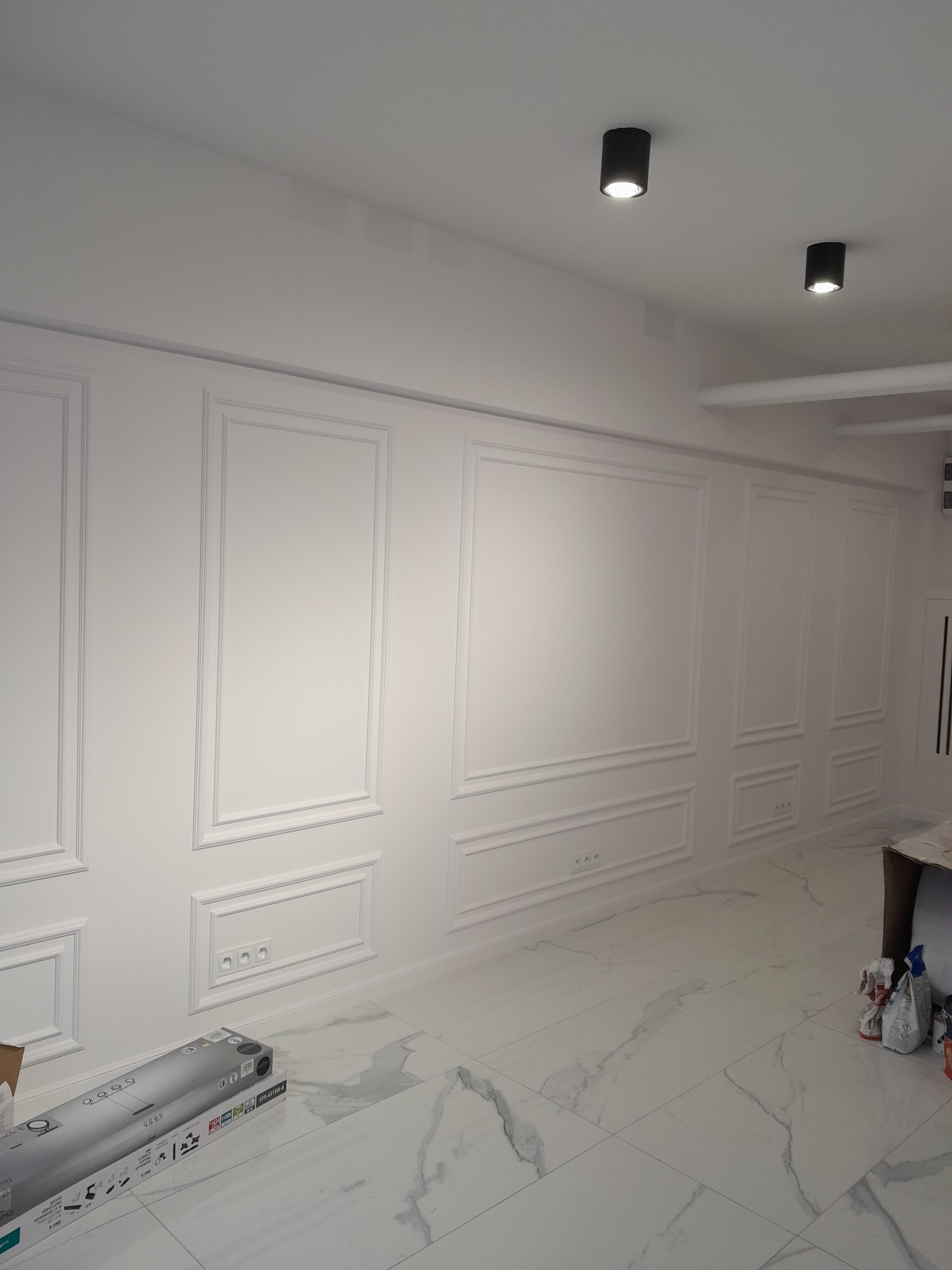 Białe ściany z dekoracyjnymi panelami i listwami, marmurowe płytki podłogowe. Widoczne gniazdka elektryczne i czarne lampy sufitowe. Częściowo widoczne materiały budowlane.