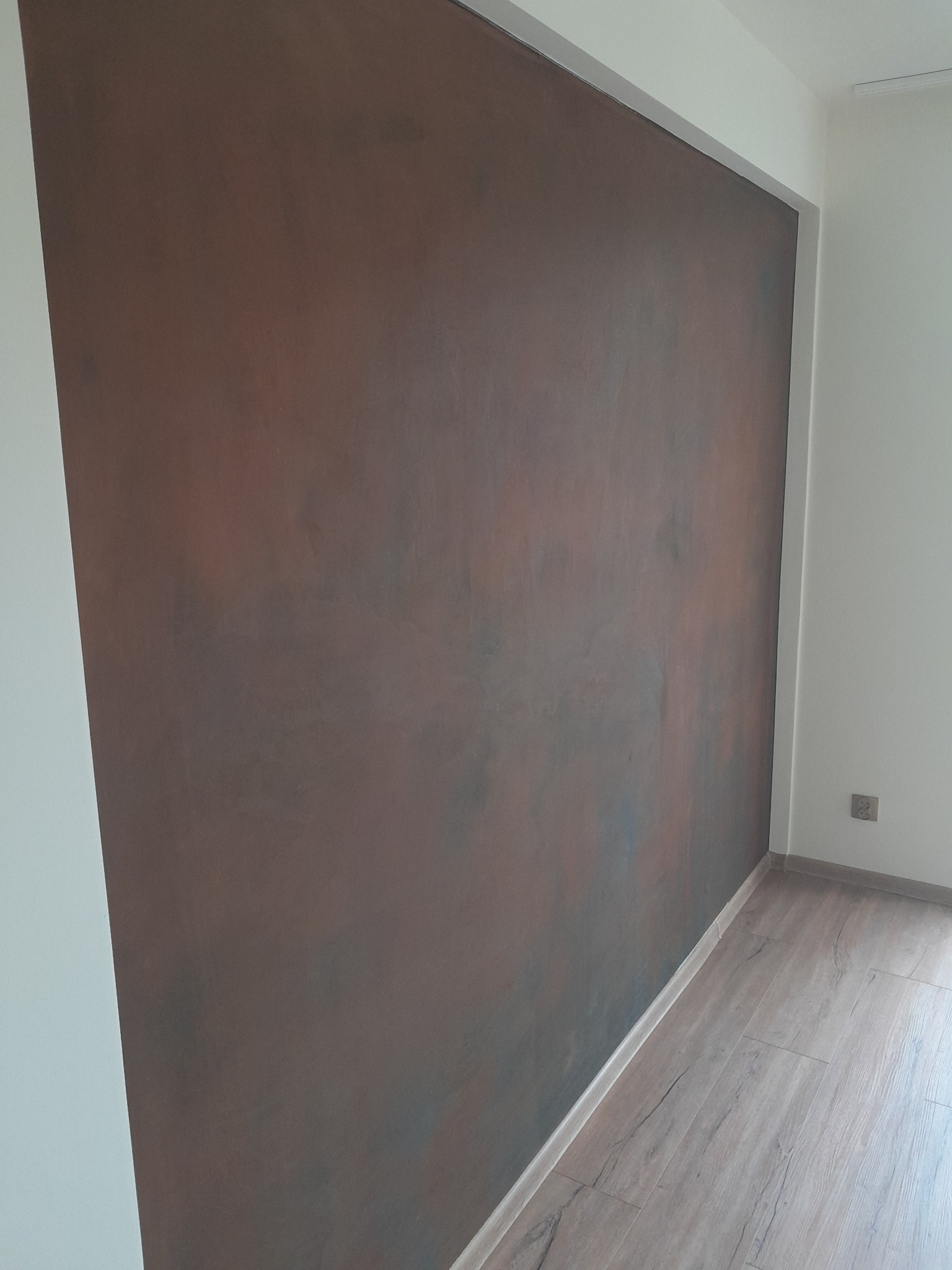 Ściana z efektem dekoracyjnym w odcieniach brązu i szarości, kontrastująca z białymi ścianami i drewnianą podłogą. Widoczny fragment gniazdka elektrycznego.
