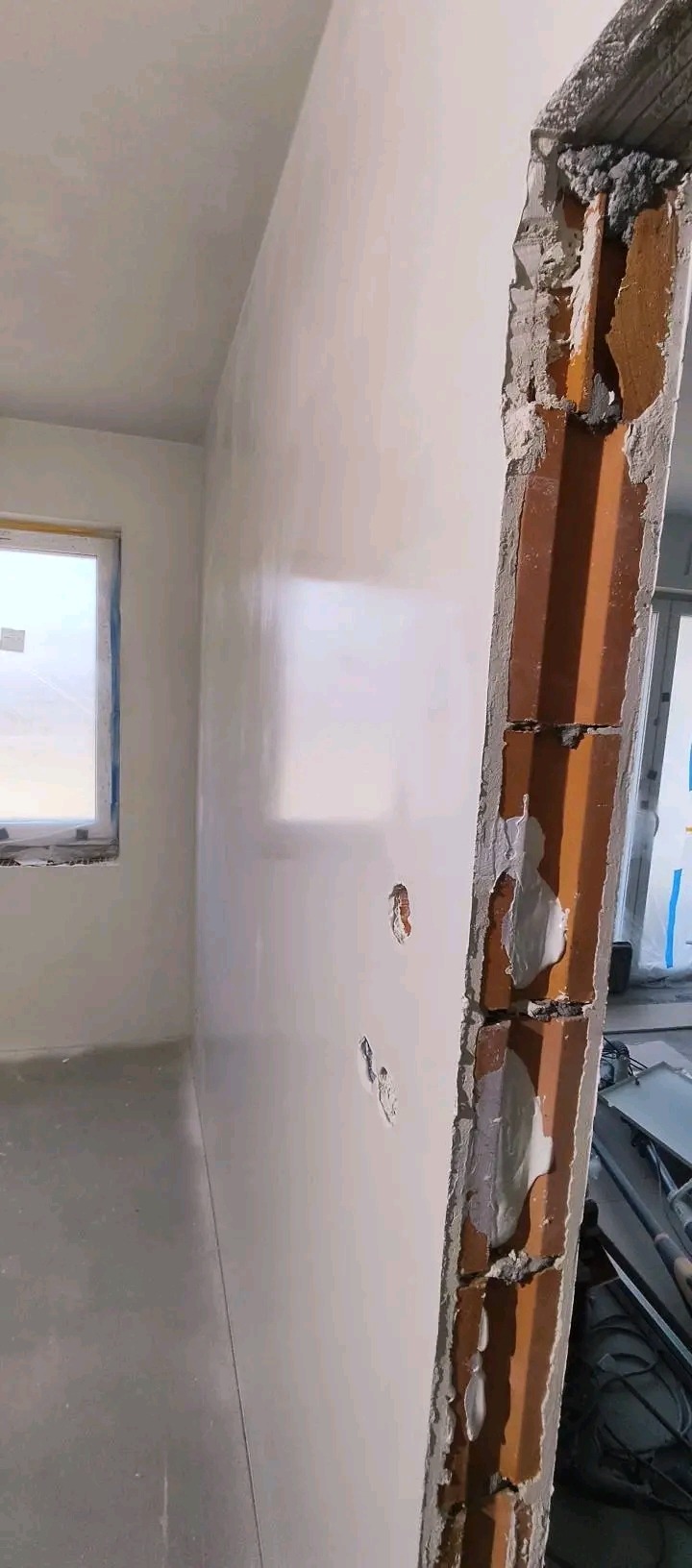 Gładka, biała ściana po szpachlowaniu w nowym wnętrzu, widoczny fragment muru z cegły i zaprawy. Okno w tle. Świeżo wykończone powierzchnie.