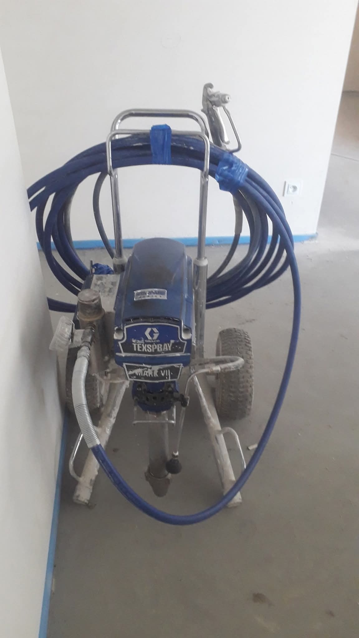 Profesjonalny agregat malarski Texspray Mark VII z niebieskim wężem, gotowy do użycia w pomieszczeniu z pomalowanymi listwami przypodłogowymi.