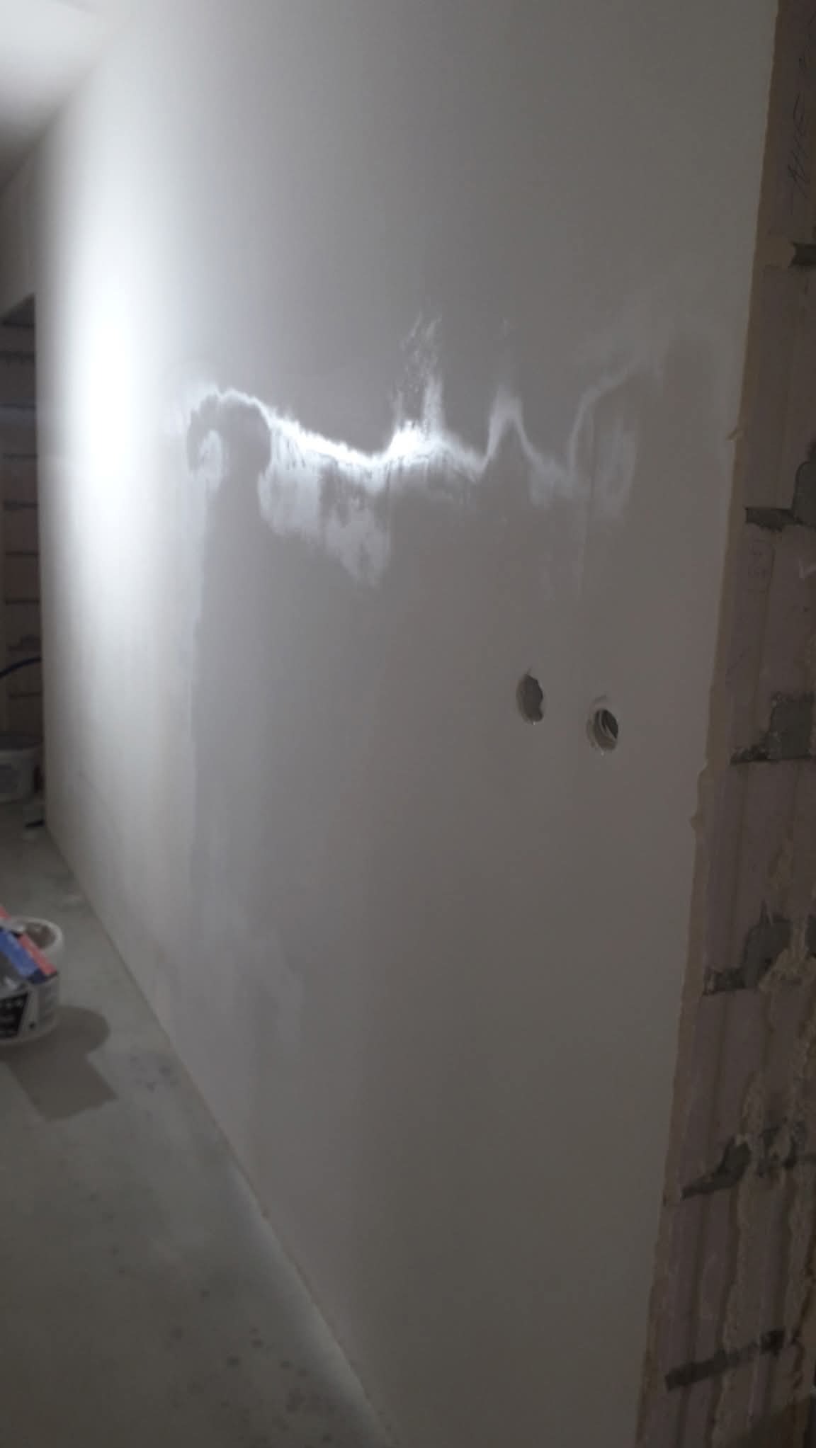 Ściana po szpachlowaniu, widoczne ślady po nałożonej gładzi. Przygotowanie do malowania, dwa otwory na gniazdka elektryczne. Fragment ściany z cegły.