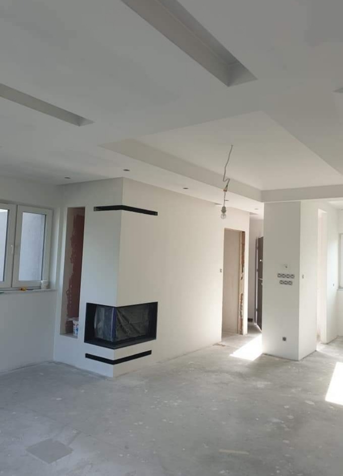 Białe wnętrze z kominkiem narożnym i oknem. Ściany i sufit pomalowane na biało, widoczne elementy dekoracyjne sufitu. Podłoga surowa, przygotowana do wykończenia.