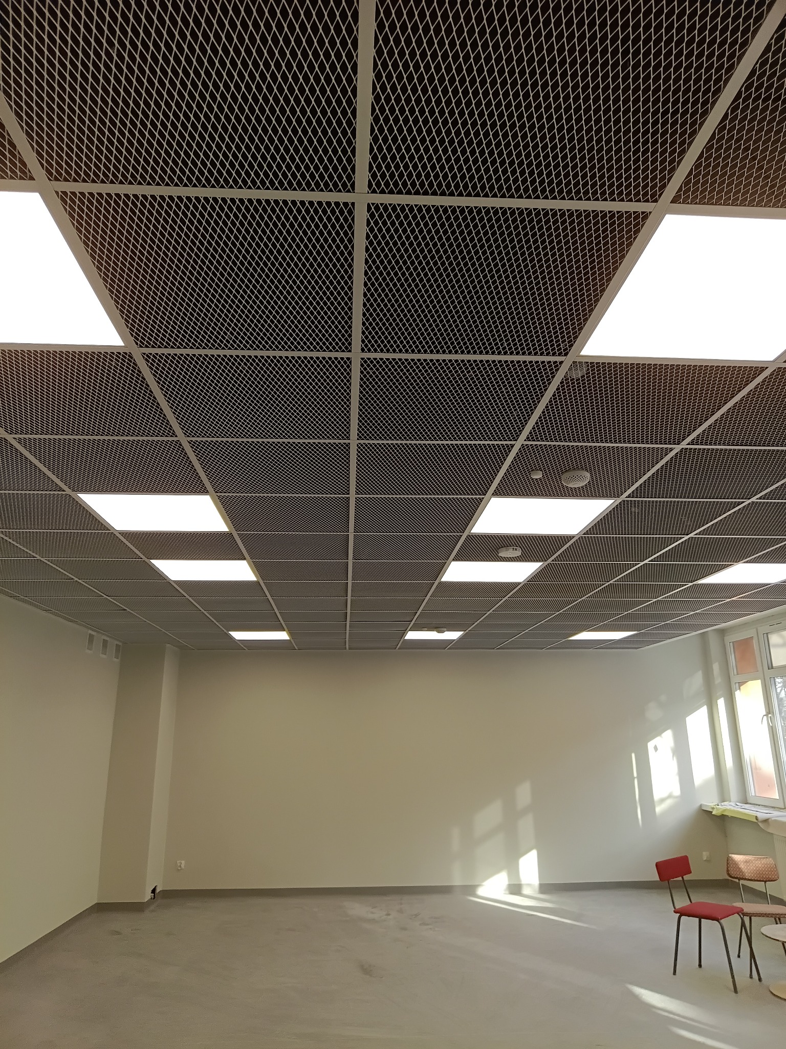 Sufit podwieszany rastrowy z oświetleniem w jasnym pomieszczeniu biurowym. Widoczne krzesła i okno z promieniami słońca. Minimalistyczny design wnętrza.