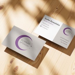 Karol Maryniak - Dwie wizytówki na drewnianym tle w ciepłym oświetleniu. Jedna z logo 'Forma Spokoju' i druga z danymi kontaktowymi studia pilatesu Sofia Kowalska.