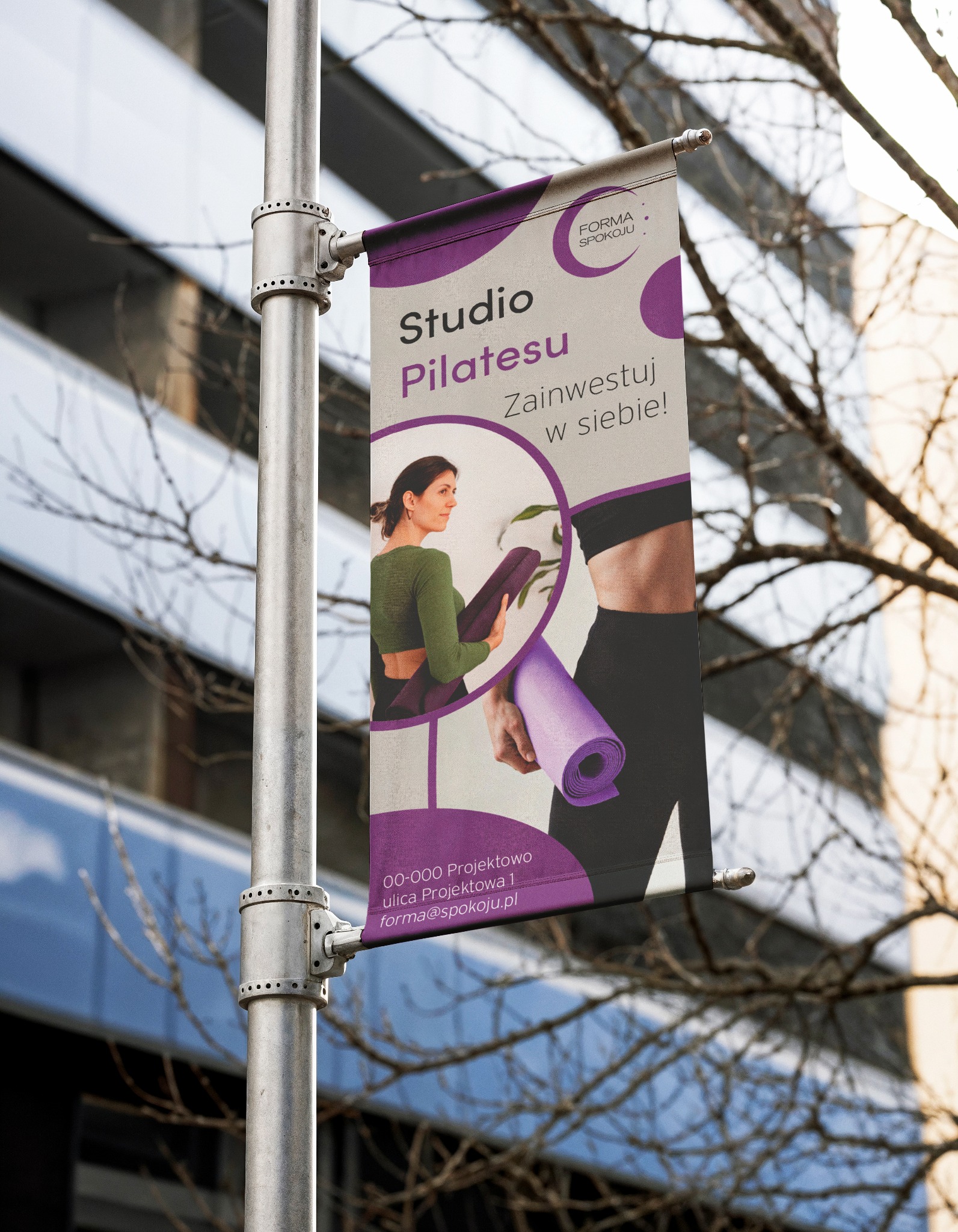 Fioletowo-biały baner reklamowy studia pilatesu 'Forma Spokoju' zawieszony na słupie oświetleniowym na tle budynku i drzewa. Baner z hasłem 'Zainwestuj w siebie!'.