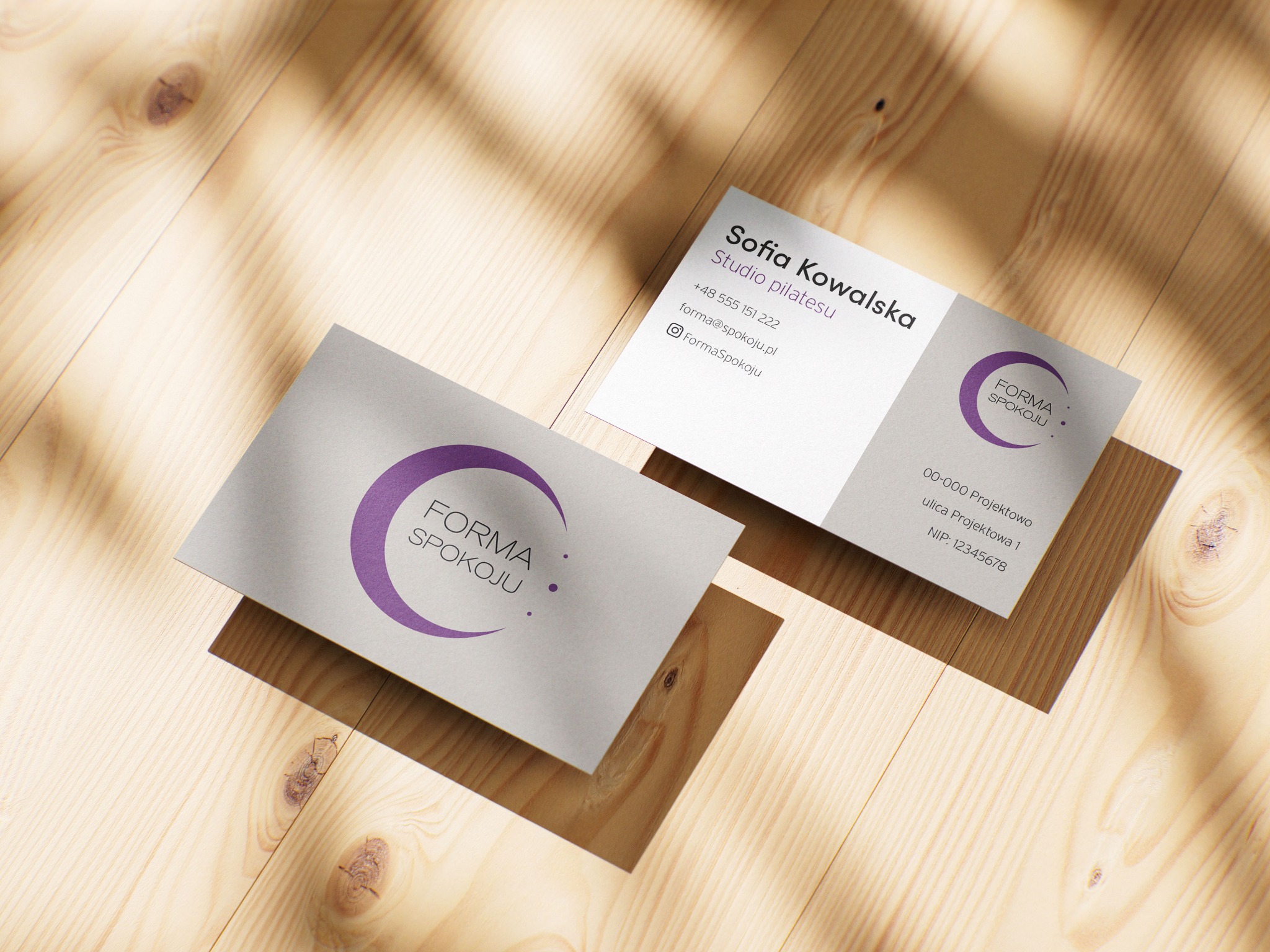 Dwie wizytówki na drewnianym tle w ciepłym oświetleniu. Jedna z logo 'Forma Spokoju' i druga z danymi kontaktowymi studia pilatesu Sofia Kowalska.