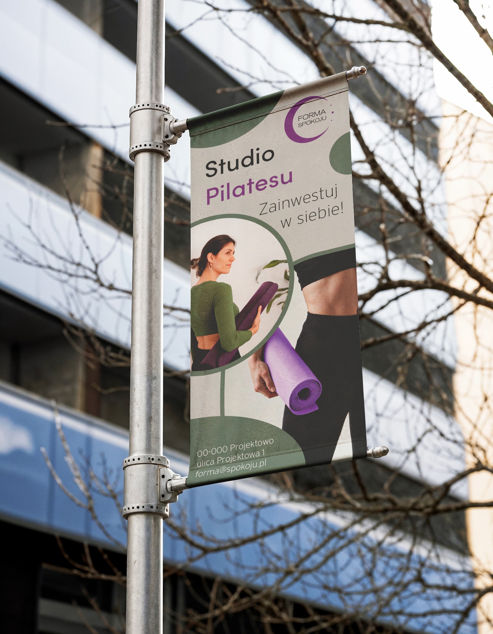 Baner reklamowy studia Pilatesu na słupie oświetleniowym, z hasłem 'Zainwestuj w siebie!' i adresem. Stonowana kolorystyka, widoczne elementy architektoniczne w tle.