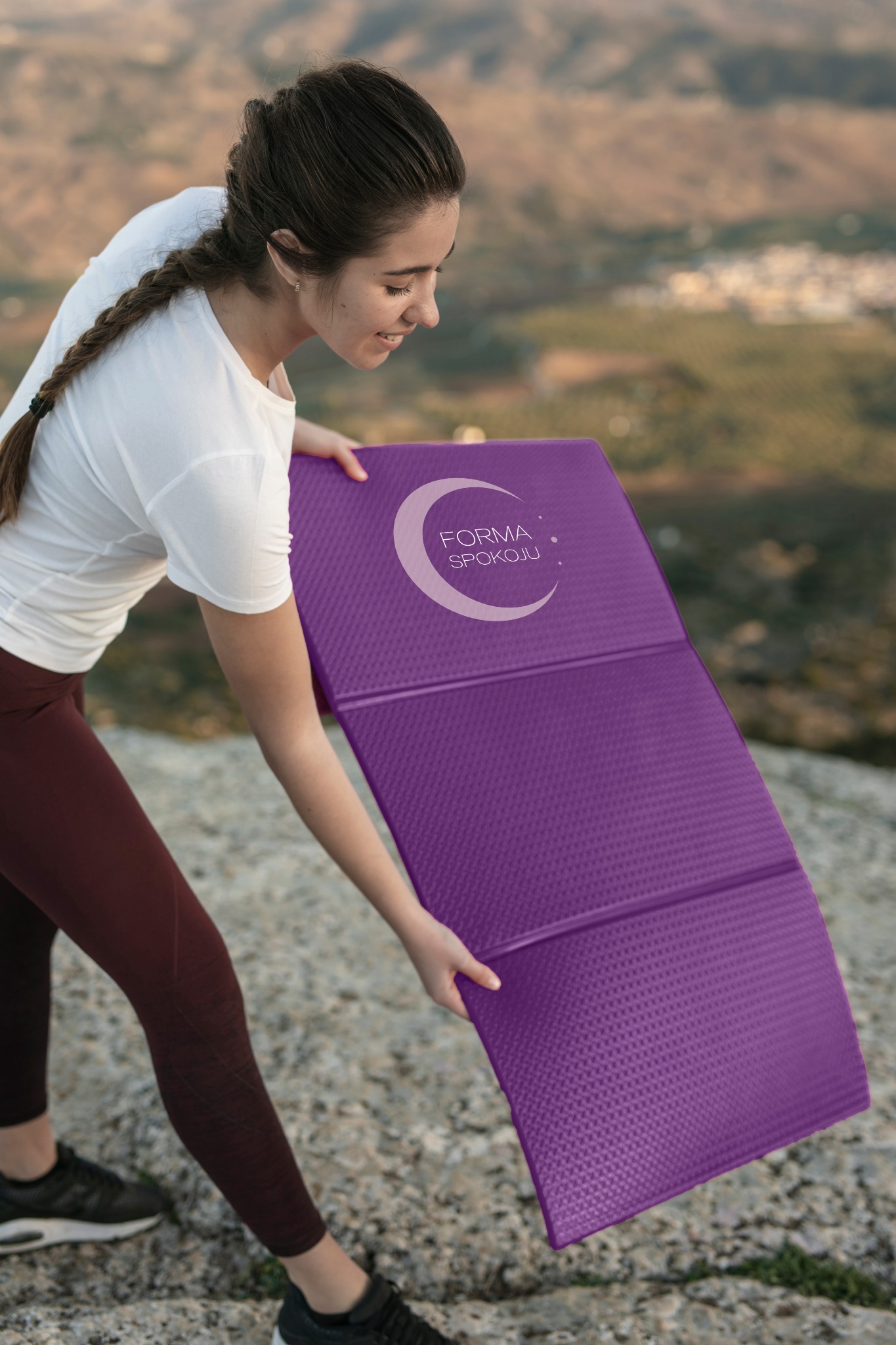 Kobieta rozkłada fioletową, składaną matę z logo 'Forma Spokoju' na tle górskiego krajobrazu. Mata ma teksturę i jest podzielona na trzy segmenty.