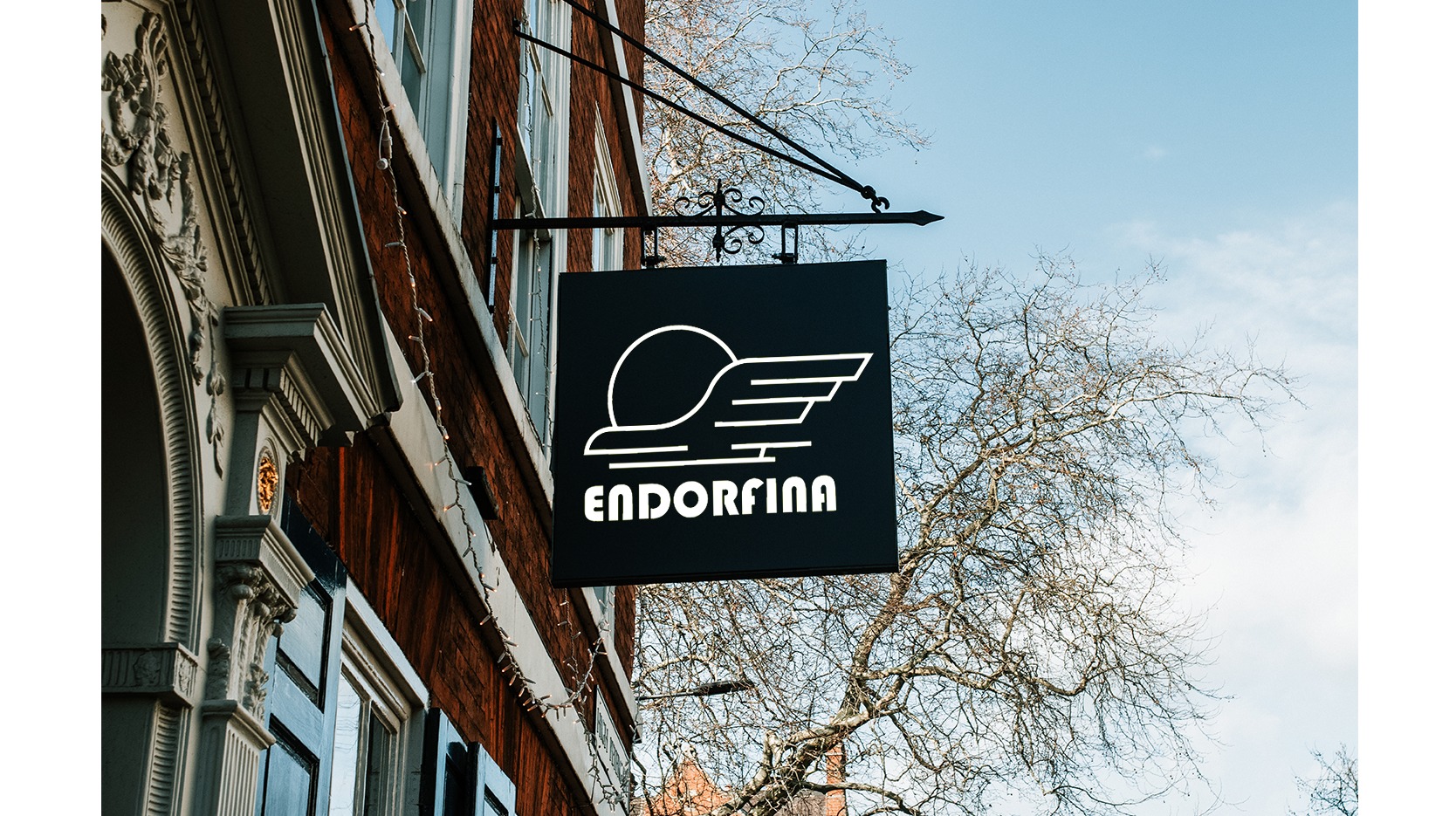 Czarny szyld kwadratowy z białym logo i nazwą 'ENDORFINA', zawieszony na ozdobnym, kutym wsporniku przy ceglanym budynku. W tle błękitne niebo i gałęzie drzewa.