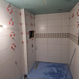 Usługi Ogólnobudowlane - Białe płytki ścienne z czerwonymi klipsami poziomującymi w trakcie układania. Fragment dekoru z motywem roślinnym. Niebieska hydroizolacja na podłodze.