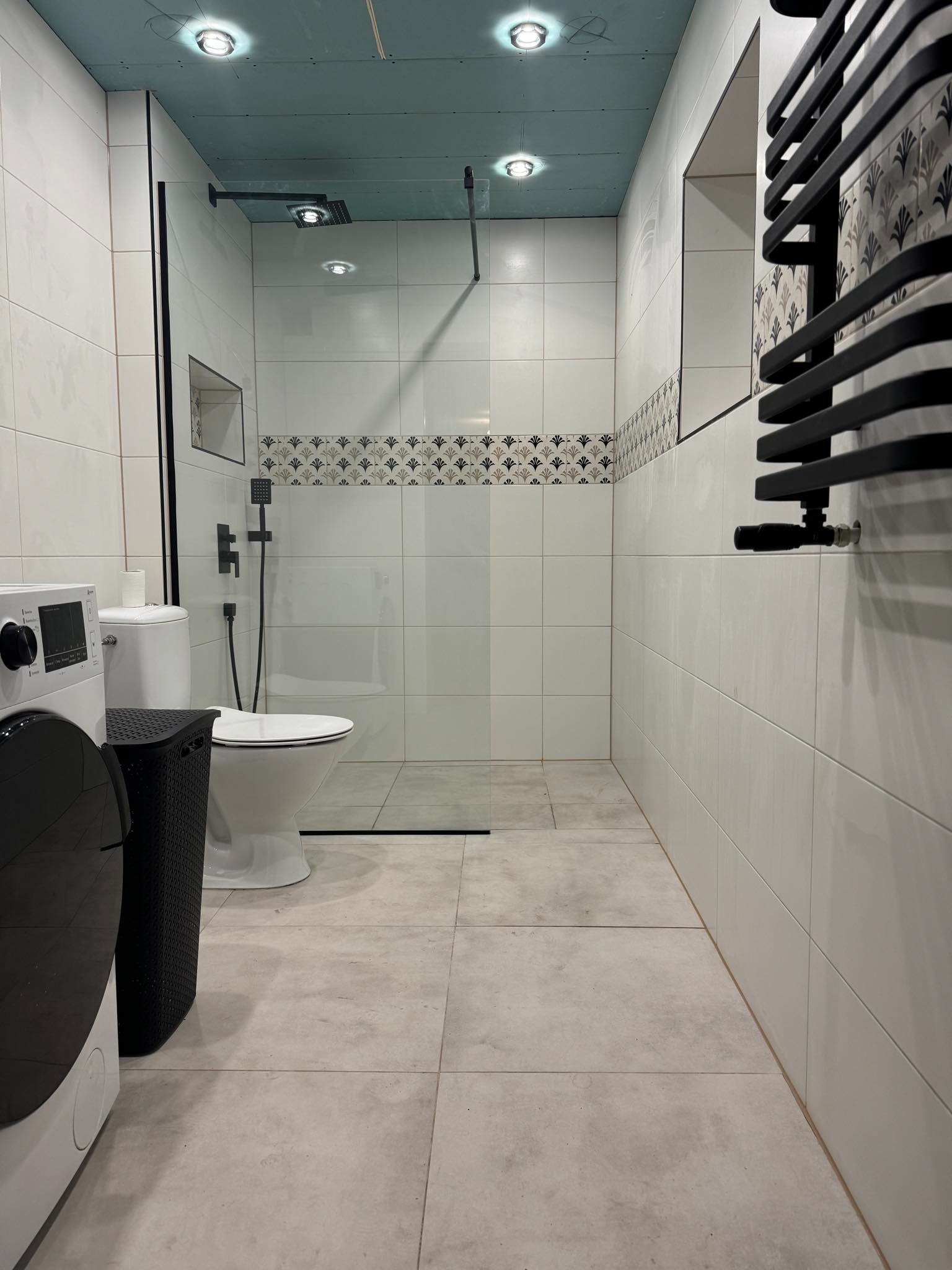 Nowoczesna łazienka z kabiną prysznicową typu walk-in, białe płytki, czarne akcenty (armatura, grzejnik), pralka z przodu. Sufit podwieszany z oświetleniem punktowym.