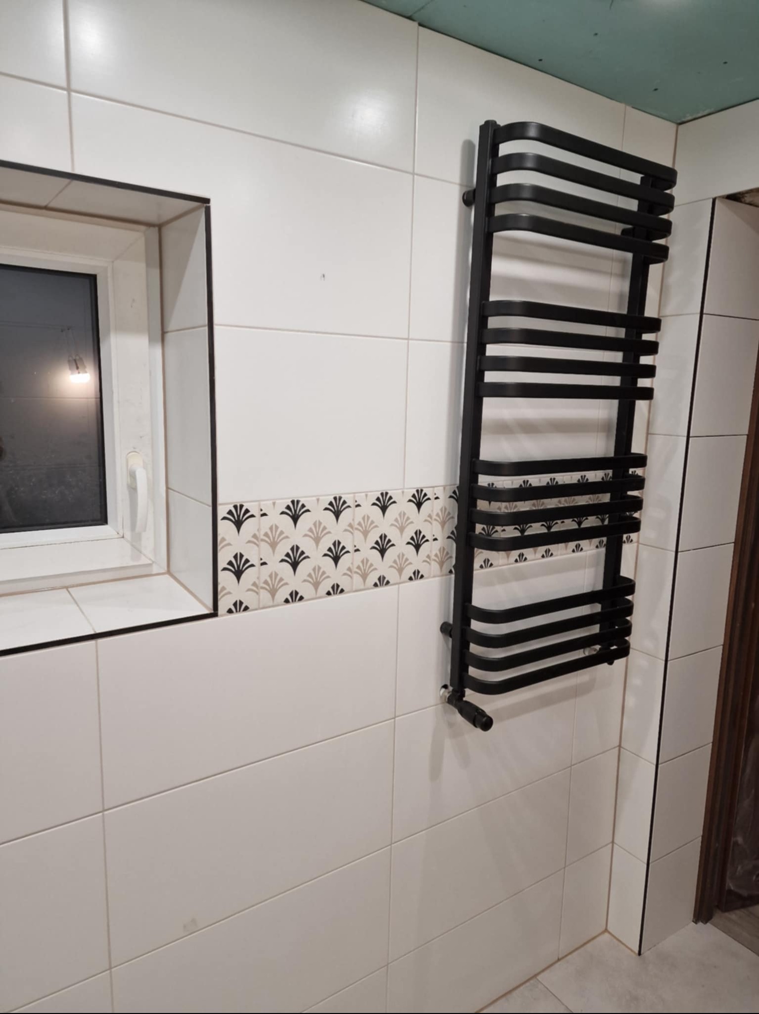 Białe płytki w łazience z dekoracyjnym pasem płytek, czarny grzejnik drabinkowy i okno z białymi ramami. Widoczny fragment sufitu z płyt karton-gips.