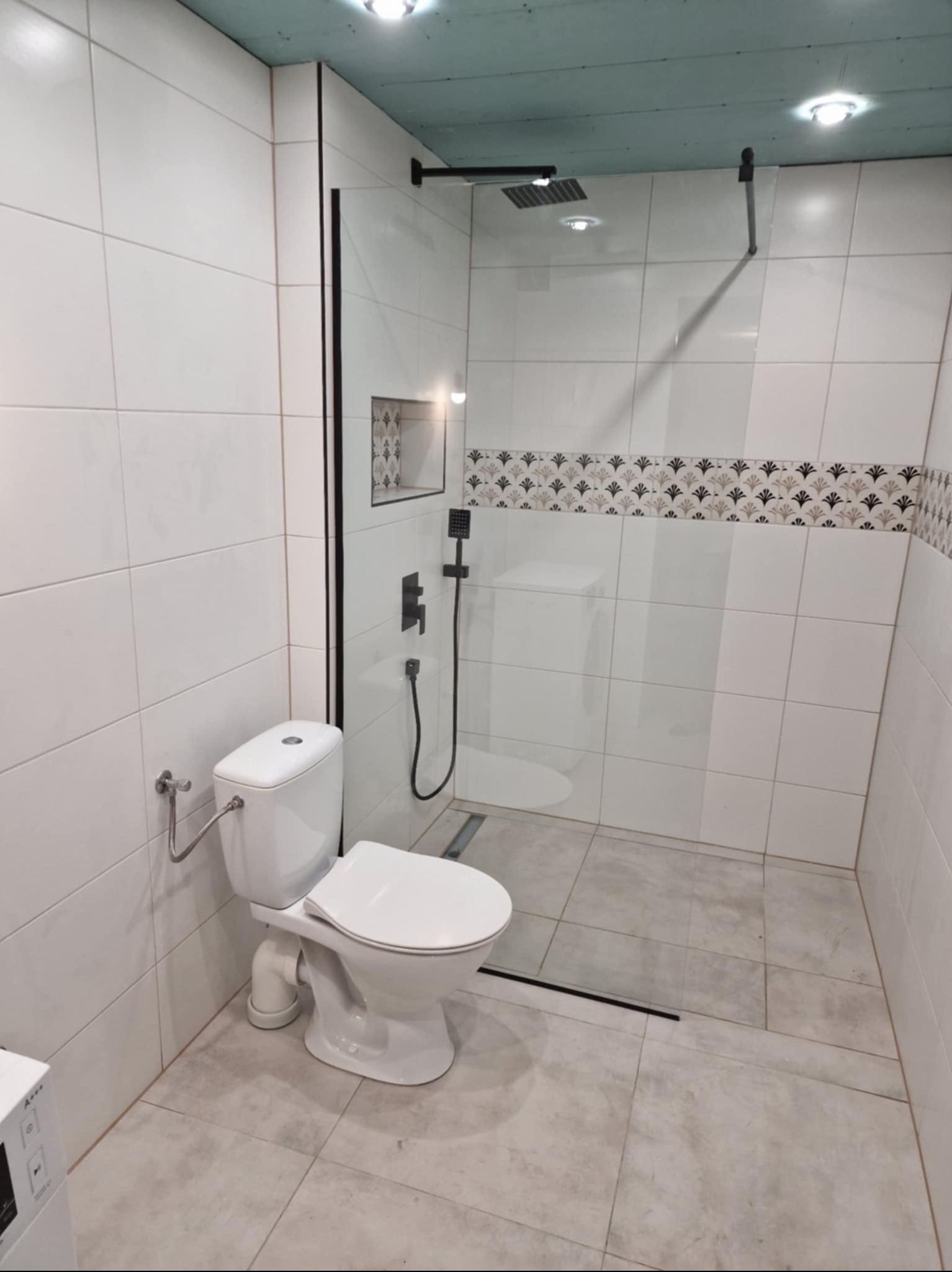 Nowoczesna łazienka z kabiną prysznicową typu walk-in, białą armaturą i dekoracyjnym pasem płytek. Minimalistyczny design z akcentami w kolorze czarnym.