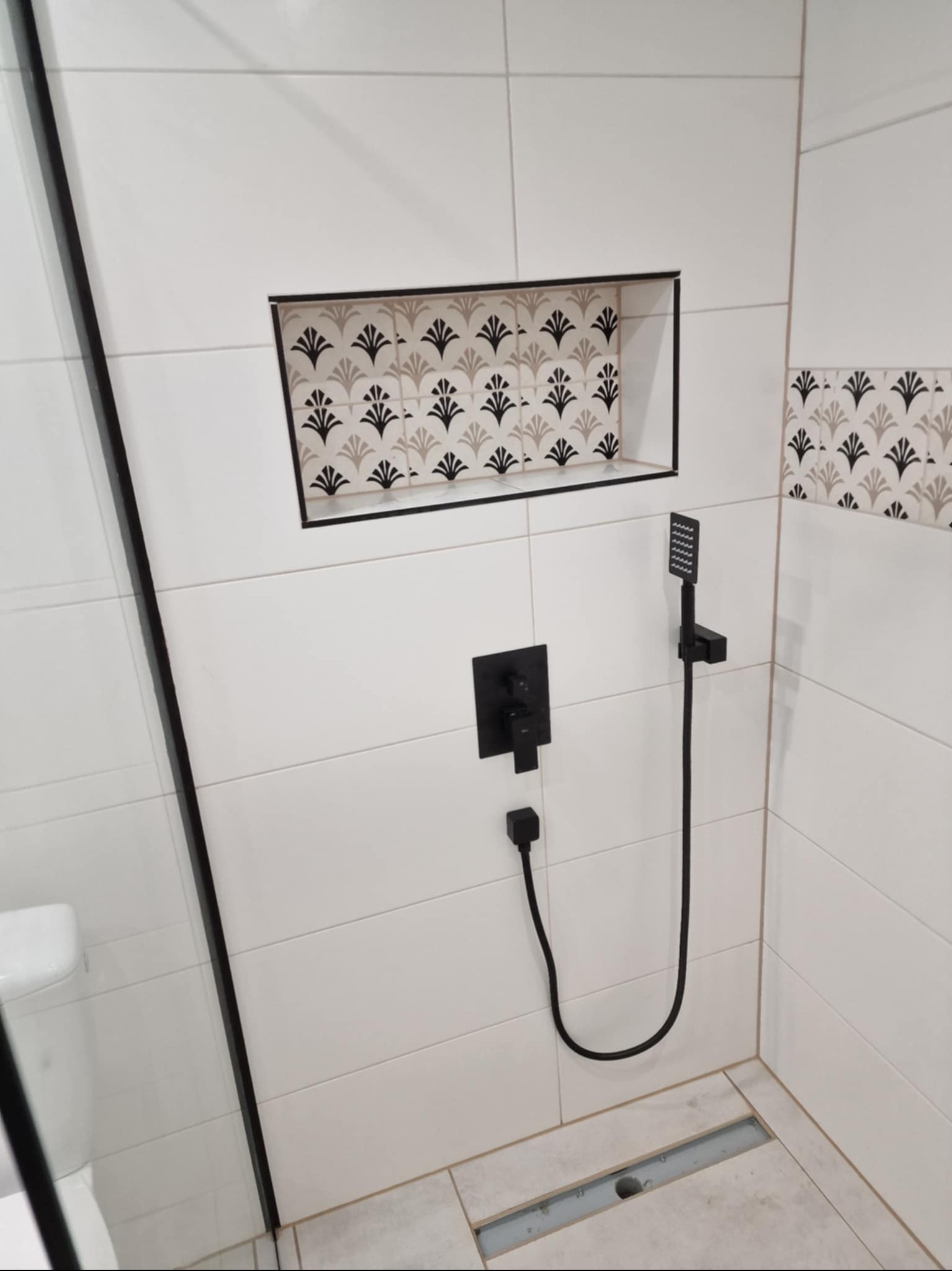 Biała łazienka z czarną armaturą i dekoracyjnymi płytkami w niszy oraz na górze ściany. Nowoczesny design z odpływem liniowym.