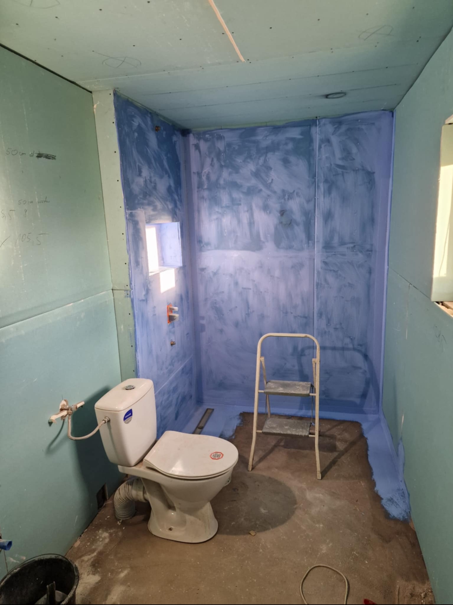 Niebieska hydroizolacja w kabinie prysznicowej z zamontowaną toaletą i drabinką. Ściany wykończone płytami gipsowo-kartonowymi z widocznymi notatkami. Etap prac wykończeniowych.