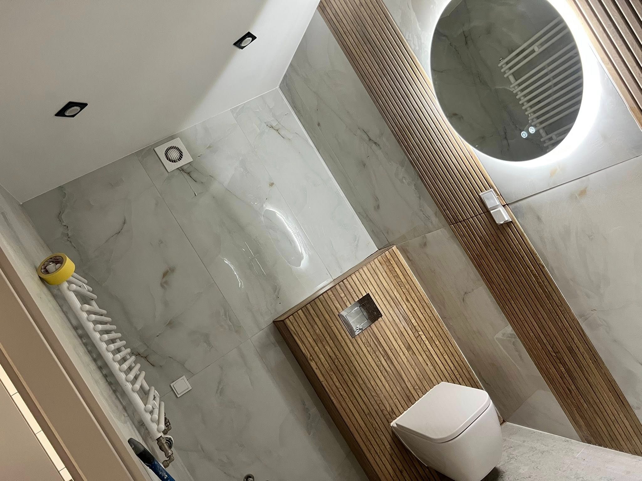 Nowoczesna łazienka po remoncie: marmurowe płytki, drewniane lamele, okrągłe lustro z podświetleniem i podwieszana toaleta. Widok z góry.